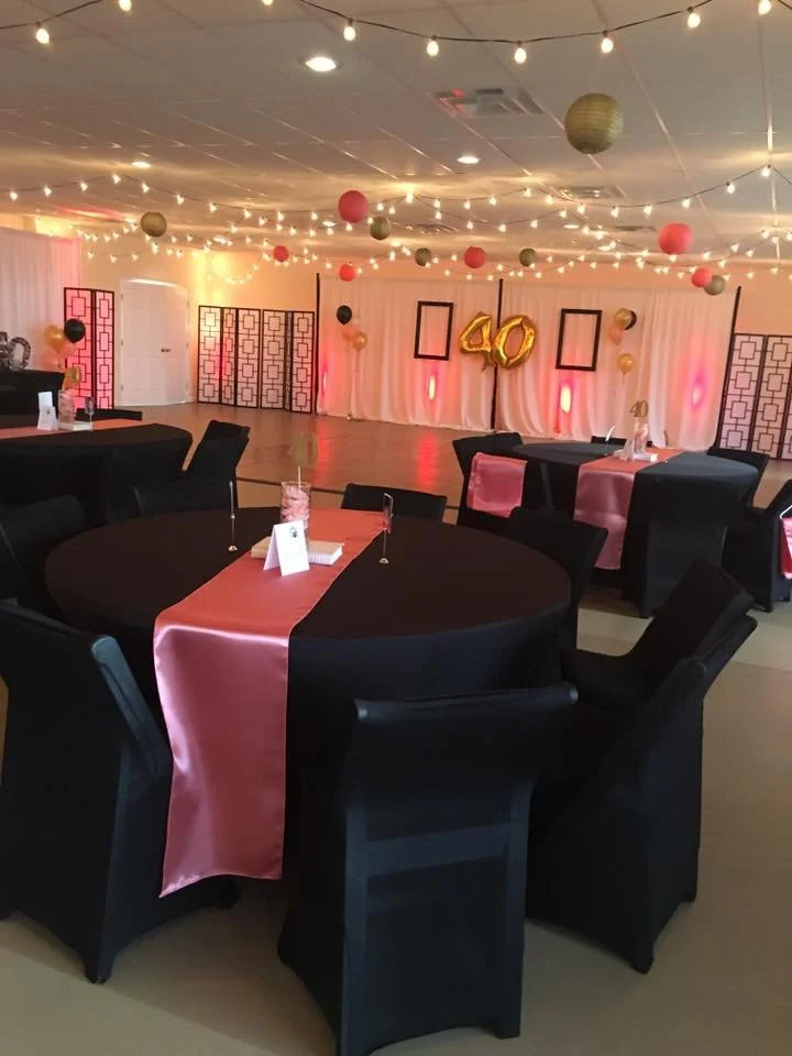 Party room decorated for a 40th birthday celebration with black tables, pink and black table runners, black chairs, and gold, pink, and black balloons. There are string lights and lanterns hanging from the ceiling, and a backdrop with gold balloons d