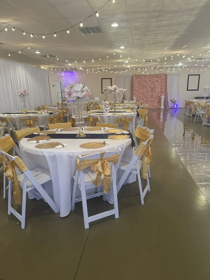Elegant event hall decorated with string lights, white drapery, and floral arrangements, featuring round tables with white tablecloths, gold seat sashes, and place settings for a celebration.