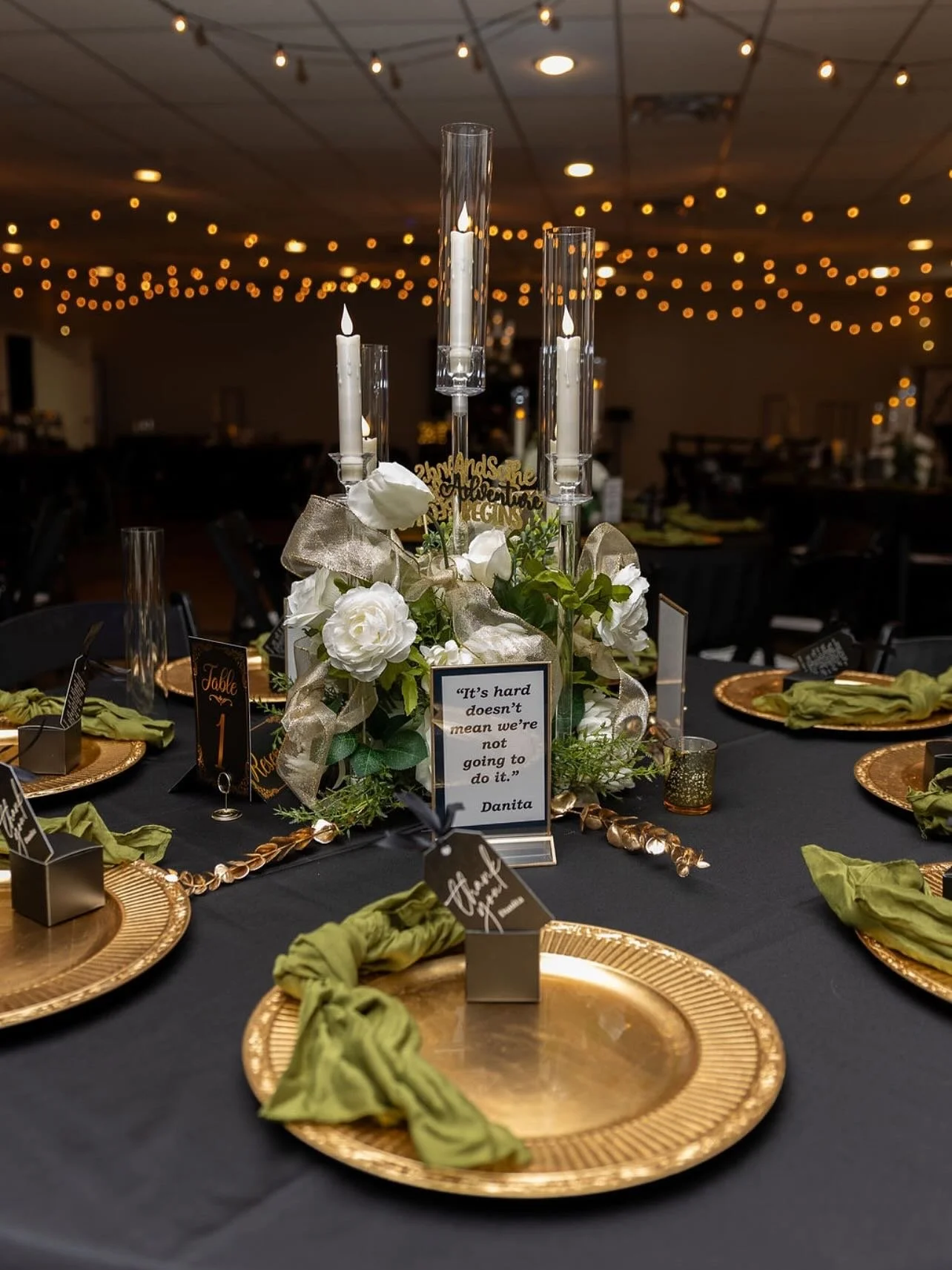 Elegant table centerpiece with white flowers, tall candles in glass holders, and gold accents, set for a formal event with gold chargers and green napkins, in a decorated hall with string lights.