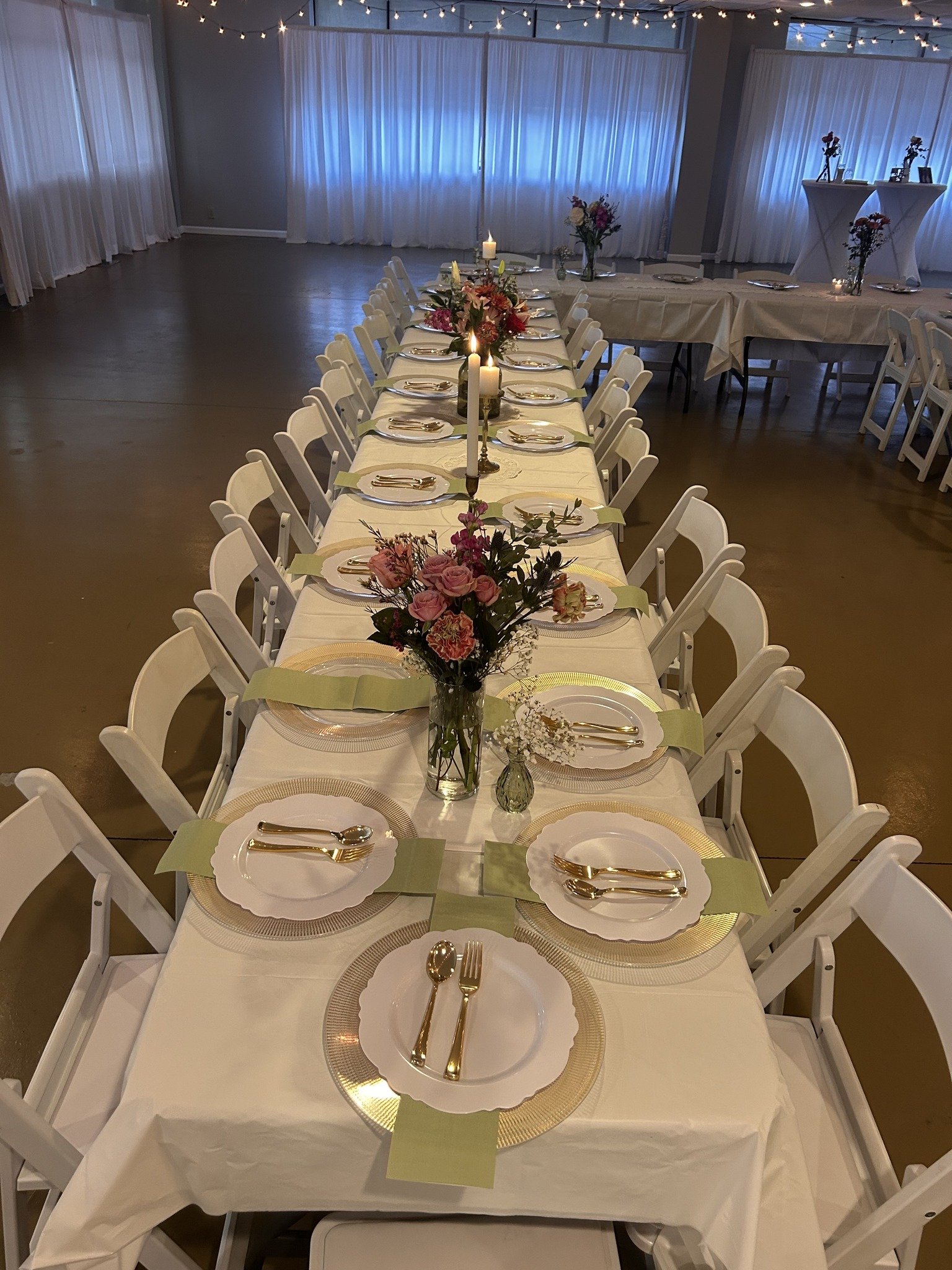 A long banquet table set for a celebration with white chairs, floral centerpieces, candles, and place settings, in a decorated room with string lights and draped curtains.