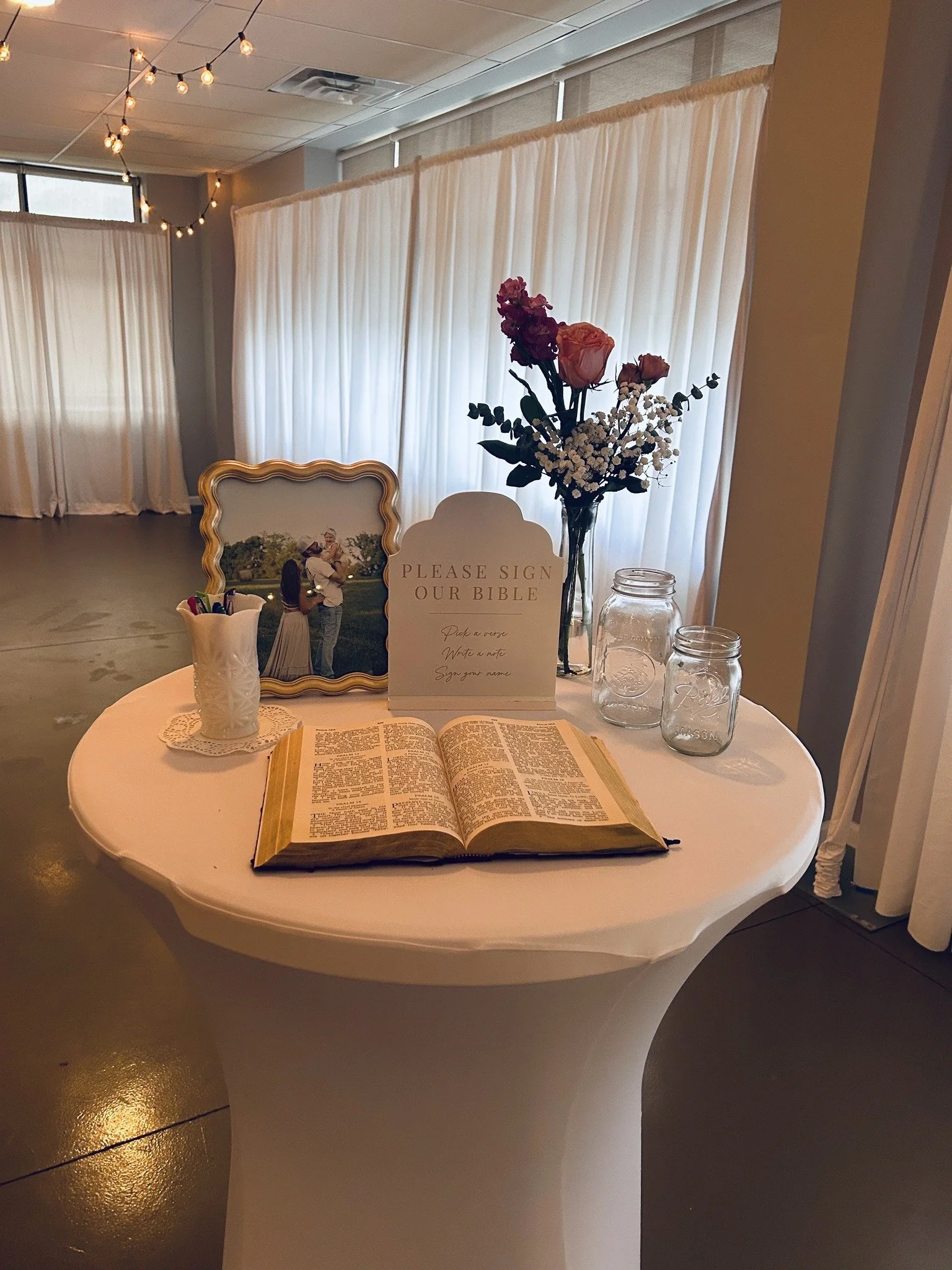 A round table with an open Bible, a framed couple's photo, a sign asking guests to sign the Bible, a vase with pink and white flowers, glass jars for collecting notes, and a cup with pens, set against white curtains and string lights in the backgroun