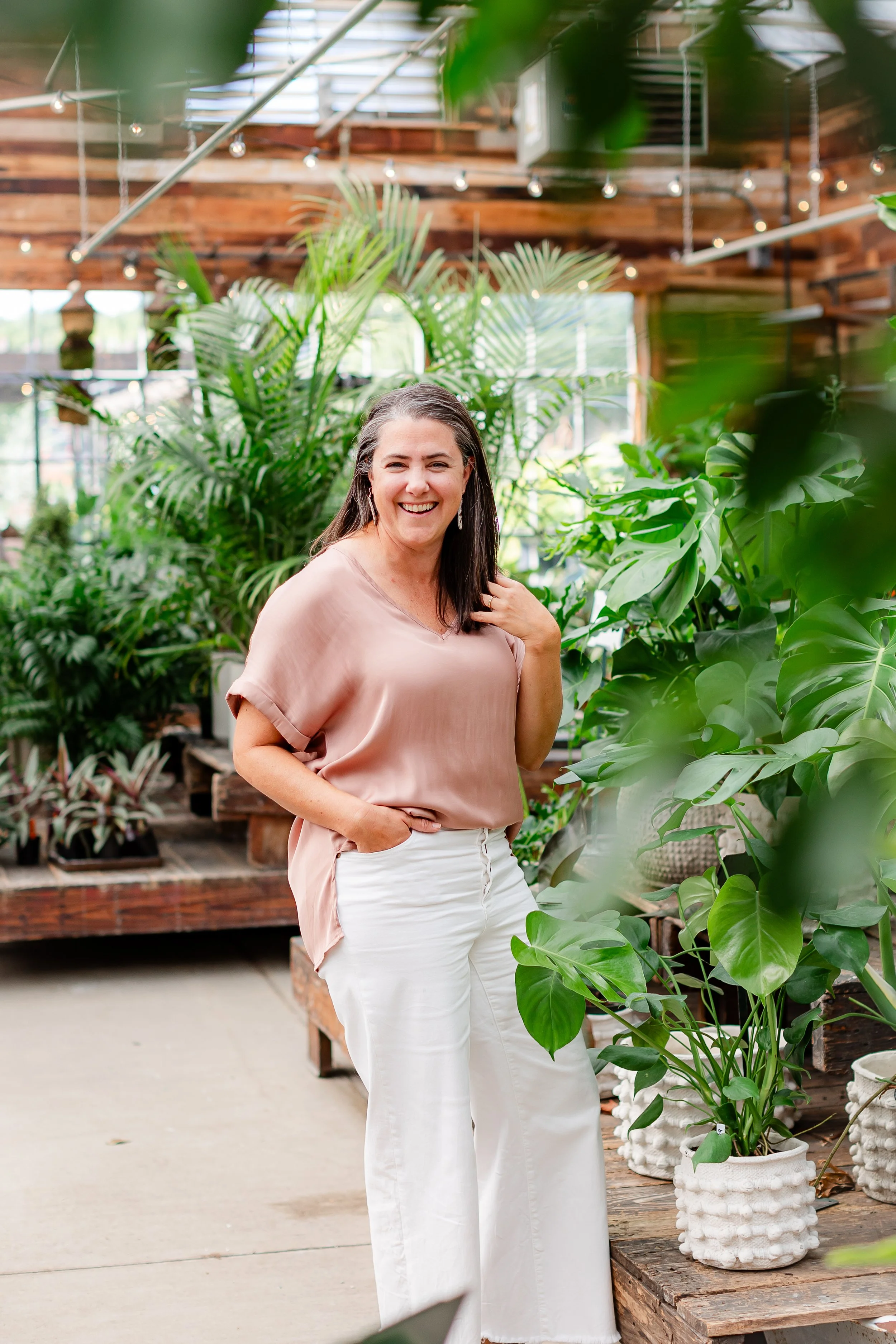A woman smiling and standing among various green plants inside a plant shop with wooden accents and natural light.