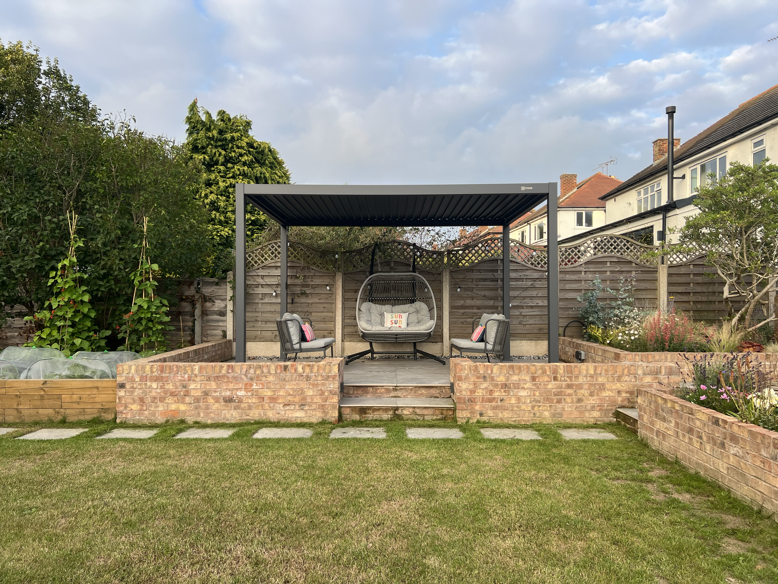 A backyard with a patio area featuring a black metal pergola, two chairs with cushions, and a hanging chair with pillows. There are flower beds on both sides with various plants and flowers, a grassy lawn in the foreground, and neighboring houses and trees in the background under a partly cloudy sky.