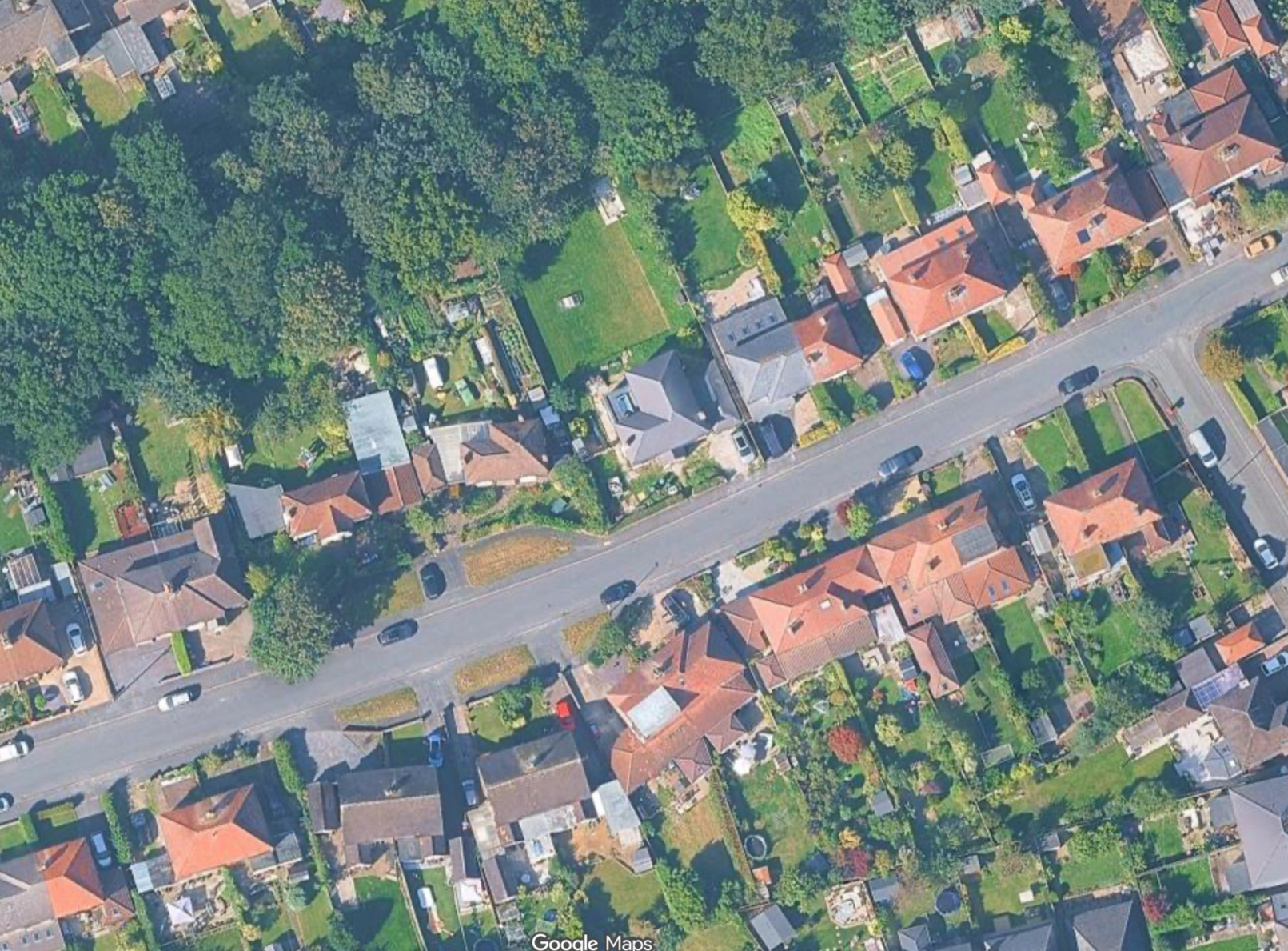 An aerial view of a suburban neighborhood showing a street with parked cars, houses with red roofs, and green yards, along with a large wooded park area to the top left.