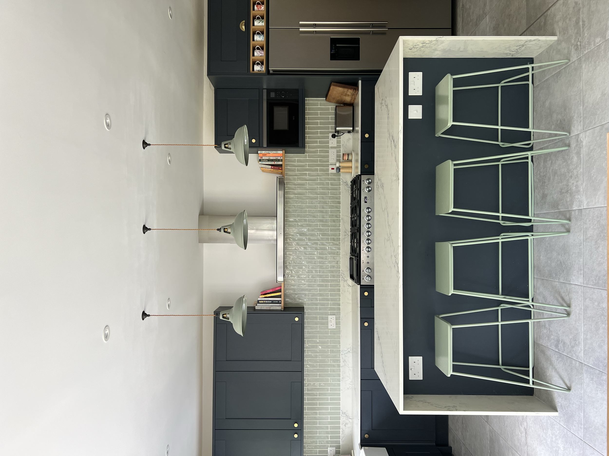 Modern kitchen with black cabinetry, a white marble island, and three green stools. Pendant lights hang above the island. The backsplash has light green tiles, and there is a black oven and microwave. Books are on the upper shelves, and outlets are on the island.