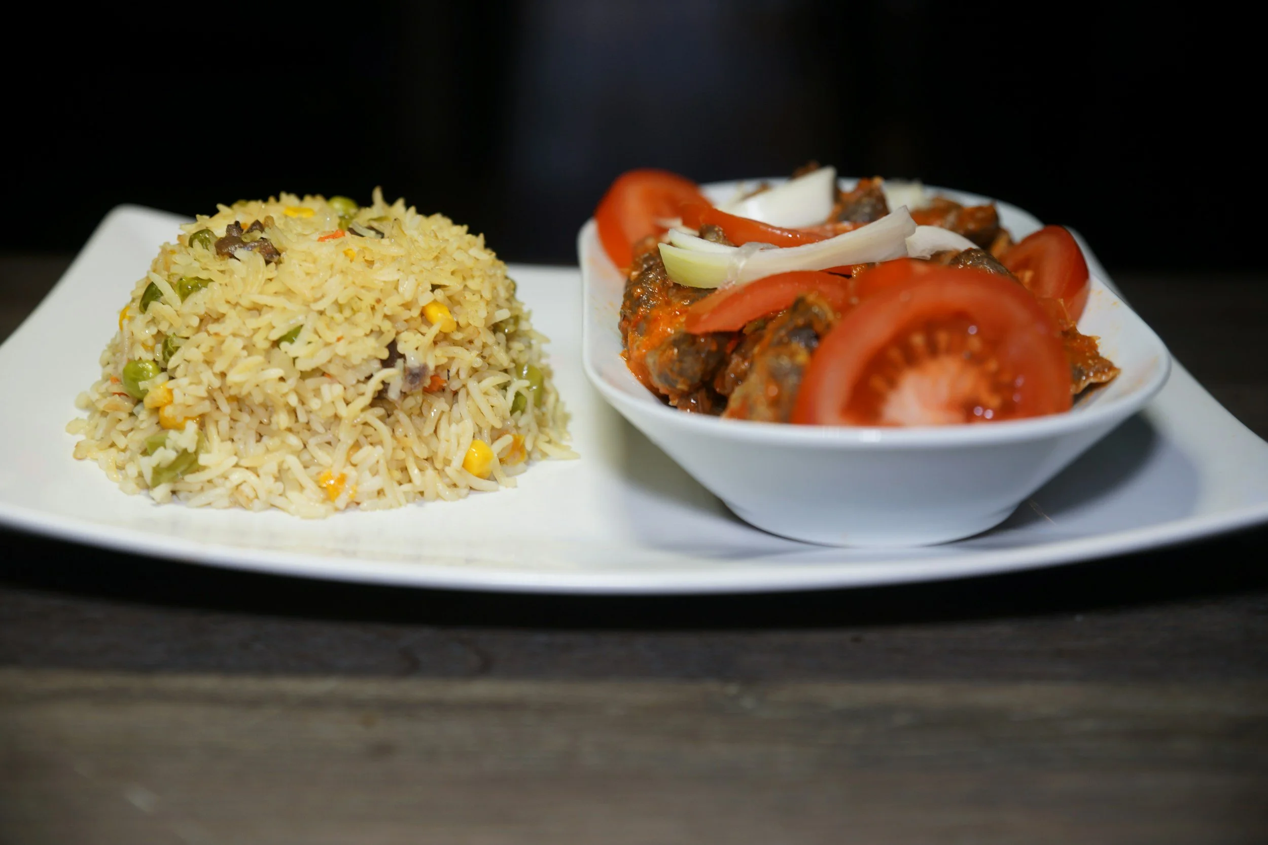 A white plate with a mound of nigerian fried rice containing vegetables such as peas, corn, and diced carrots, and a small white bowl of salad with sliced tomatoes and onions.