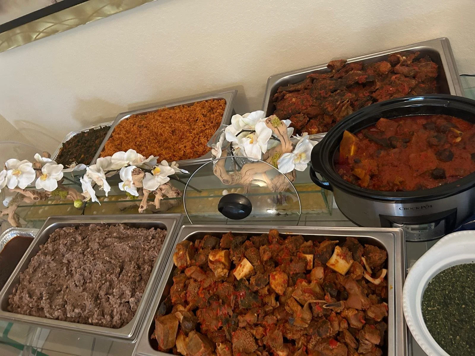 Assorted bowls and trays of assorted beef, ayamase, ewa aganyin, jollof rice, and vegetables on a table, decorated with artificial white orchids.