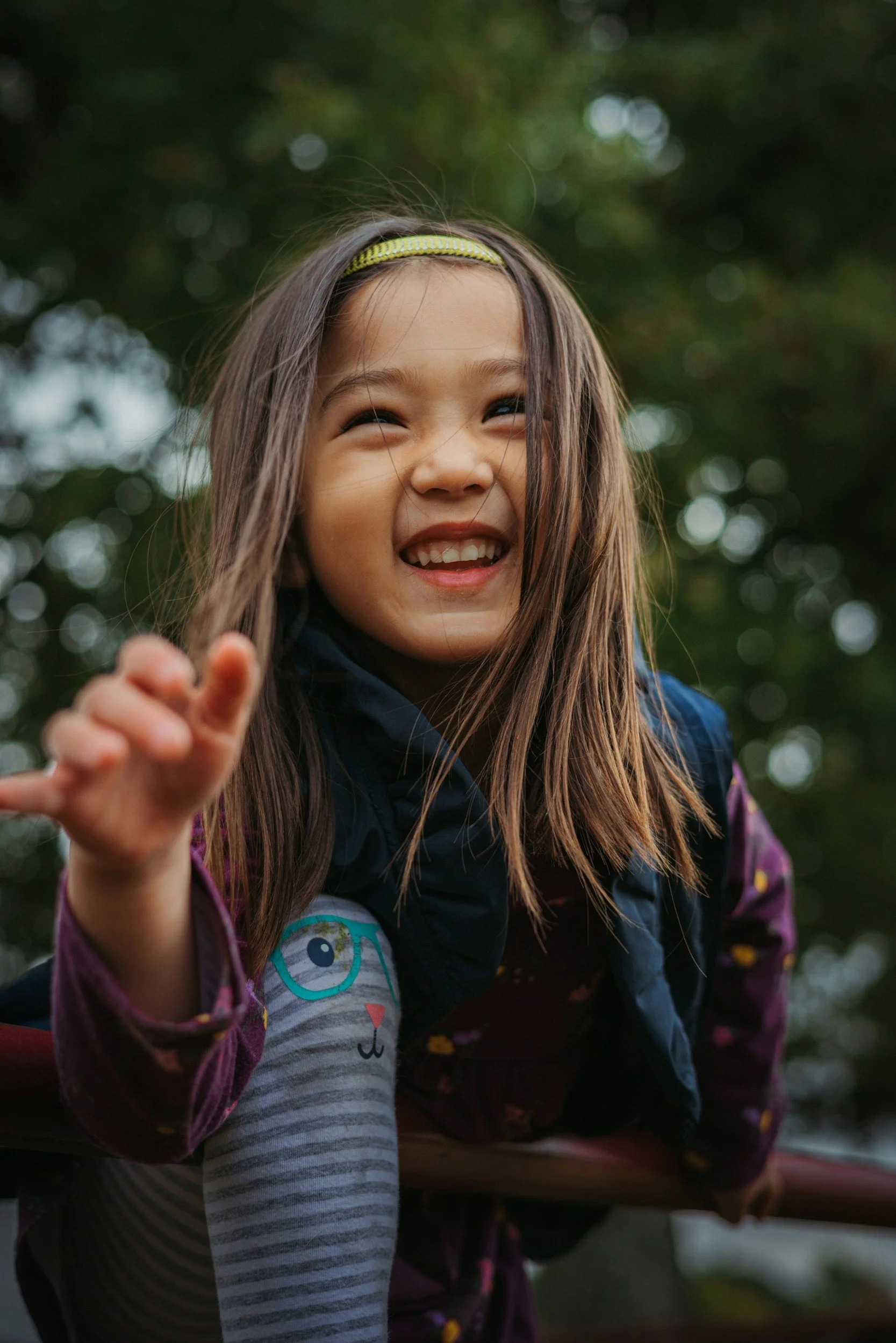 Smiling young girl with long hair, wearing a headband, dark blue jacket, and striped leggings with a cat face design, reaching out her hand outdoors against a blurry background of trees.
