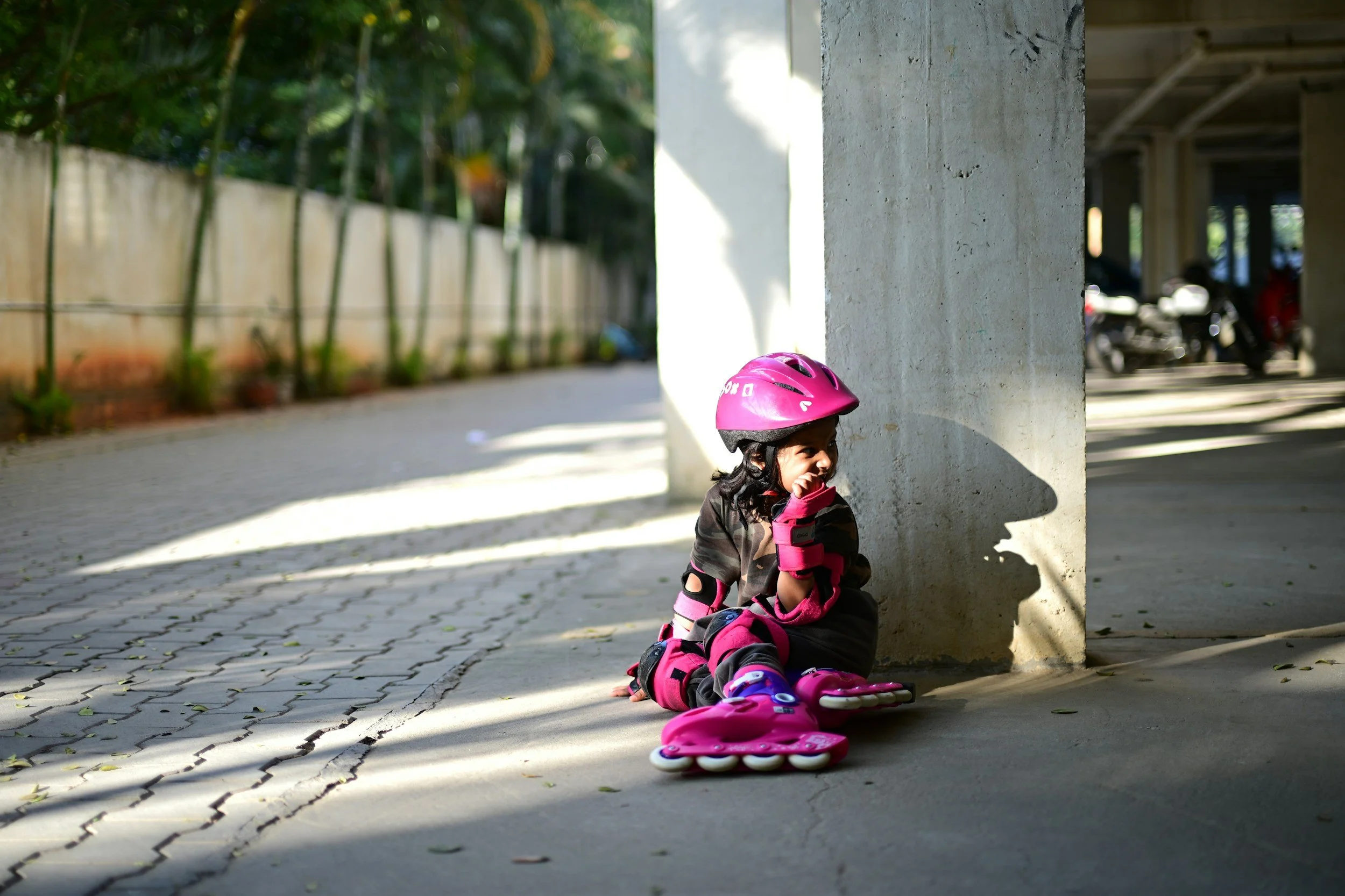 A young girl wearing pink rollerblades, a pink helmet, and protective gear, sitting on the ground under a concrete structure, smiling while touching her face. She is outdoor with sunshine and shadows on the pavement.