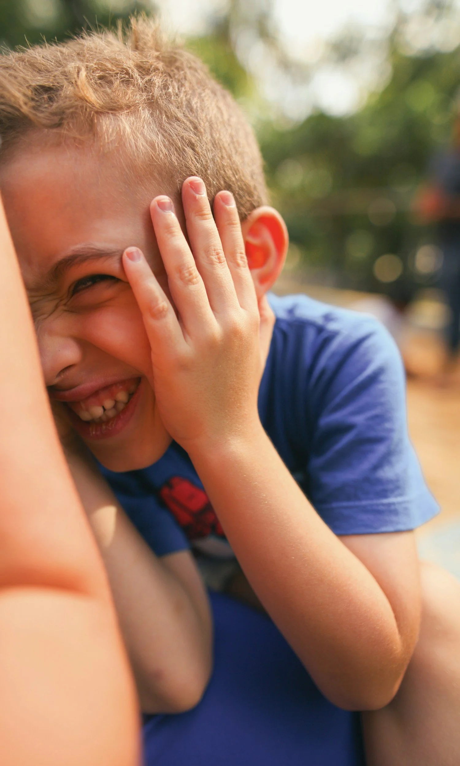 A young boy with short blond hair covering his face with his hand, laughing.