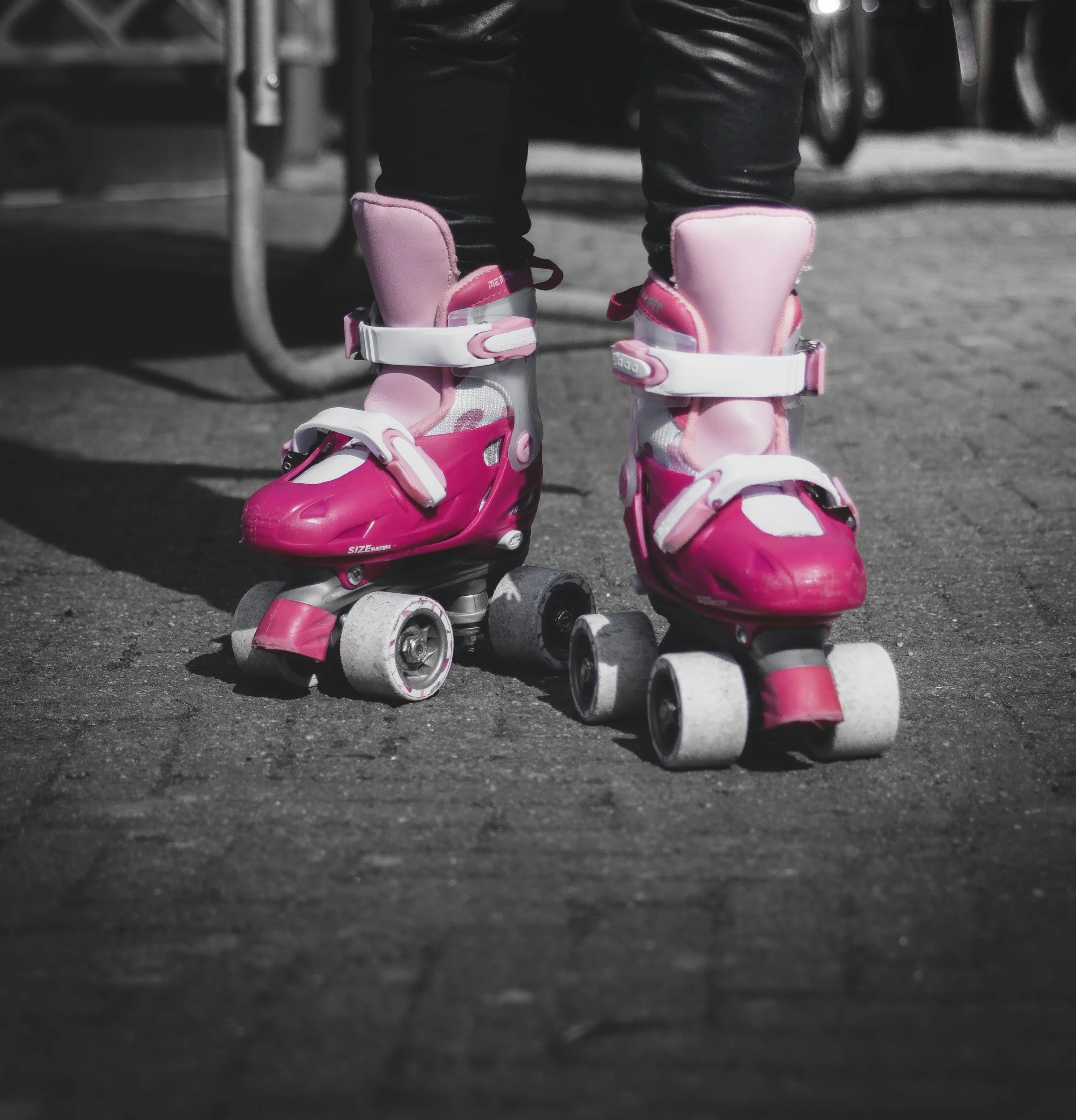 Pink inline skate with white straps resting on a black asphalt surface, with black pants visible above the skate blades.