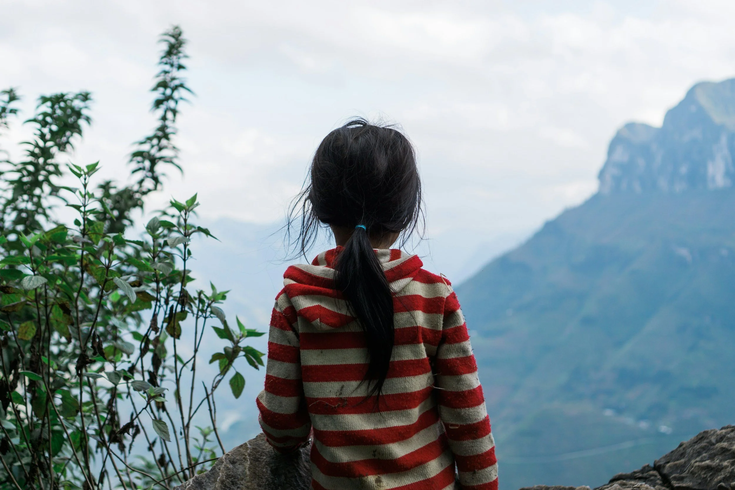 A girl with long dark hair tied back in a ponytail, wearing a red and beige striped sweater, sitting on a rock and looking at a mountainous landscape with cloudy sky.