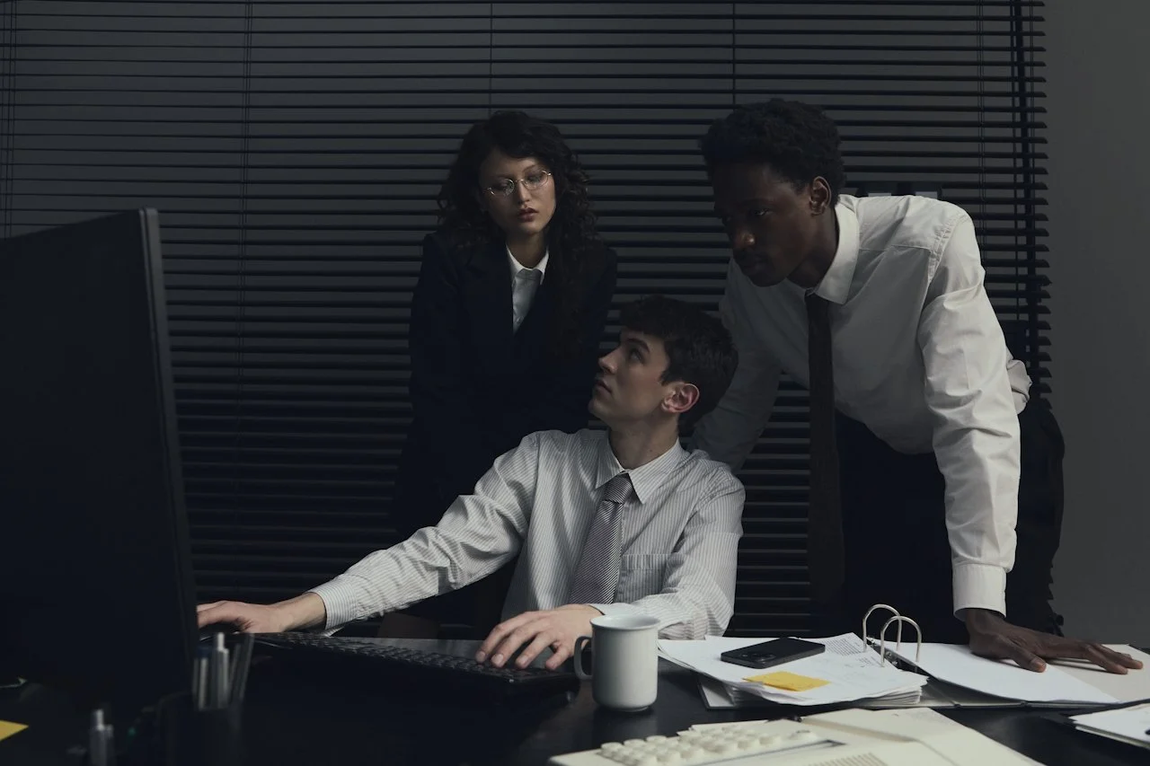 Three businesspeople in an office, two men and one woman, looking at a computer screen with serious expressions.