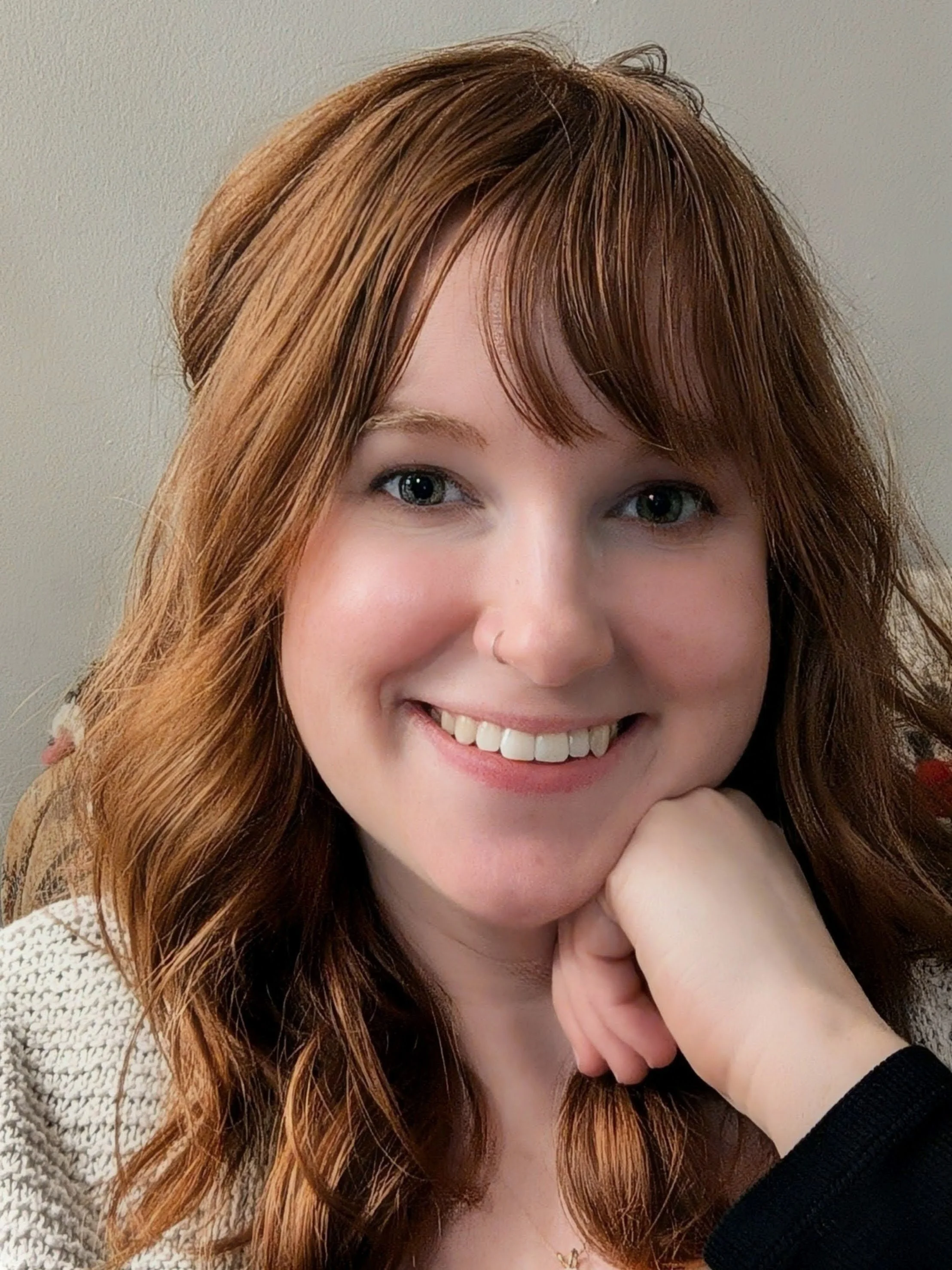 Close-up of a smiling woman therapist with red hair, green eyes, and a nose ring, resting her chin on her hand.