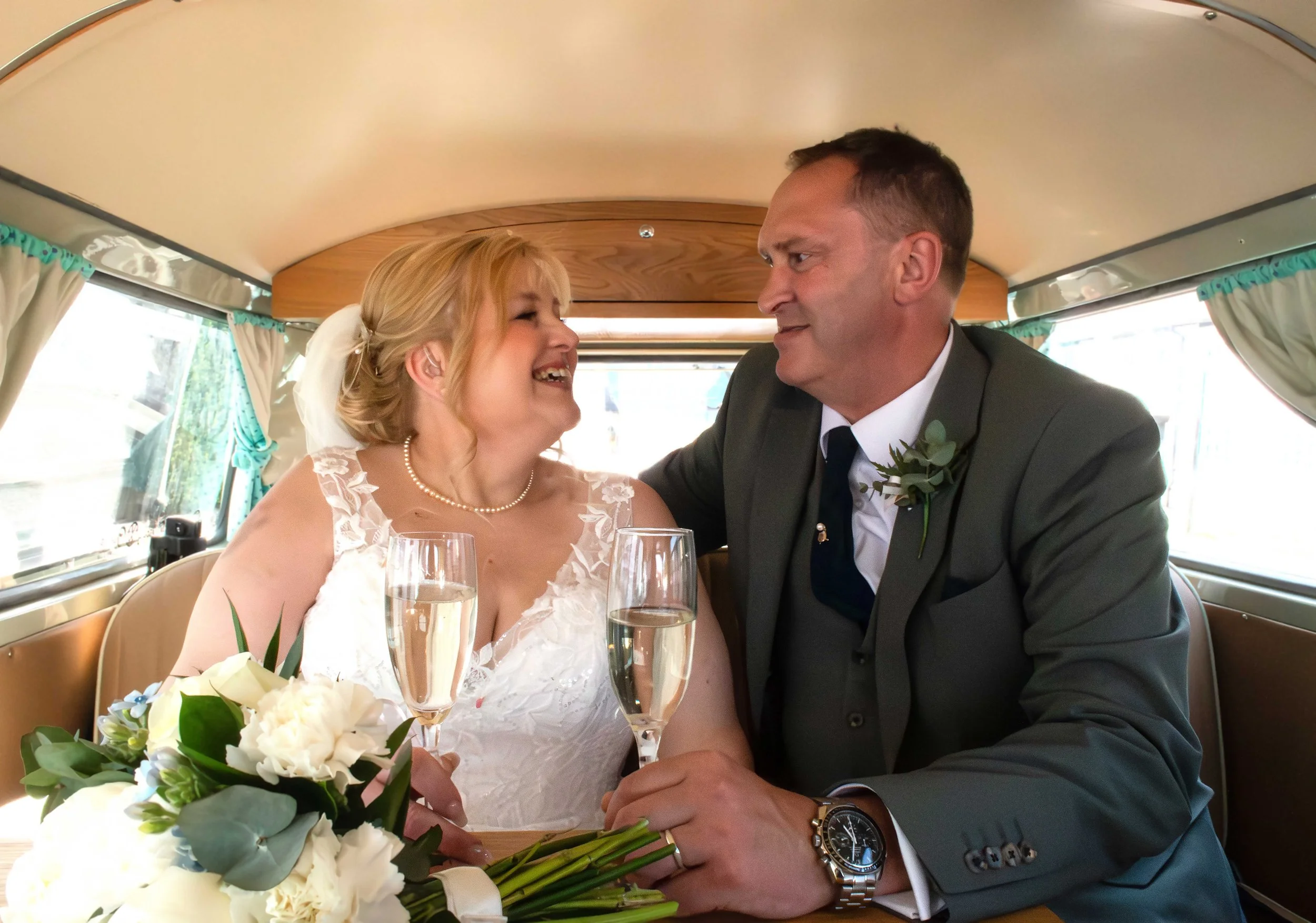 A bride and groom sitting together in a vehicle, smiling and looking at each other, holding glasses of champagne, with a bouquet of flowers on the table at the St Andrews Church, Headington
