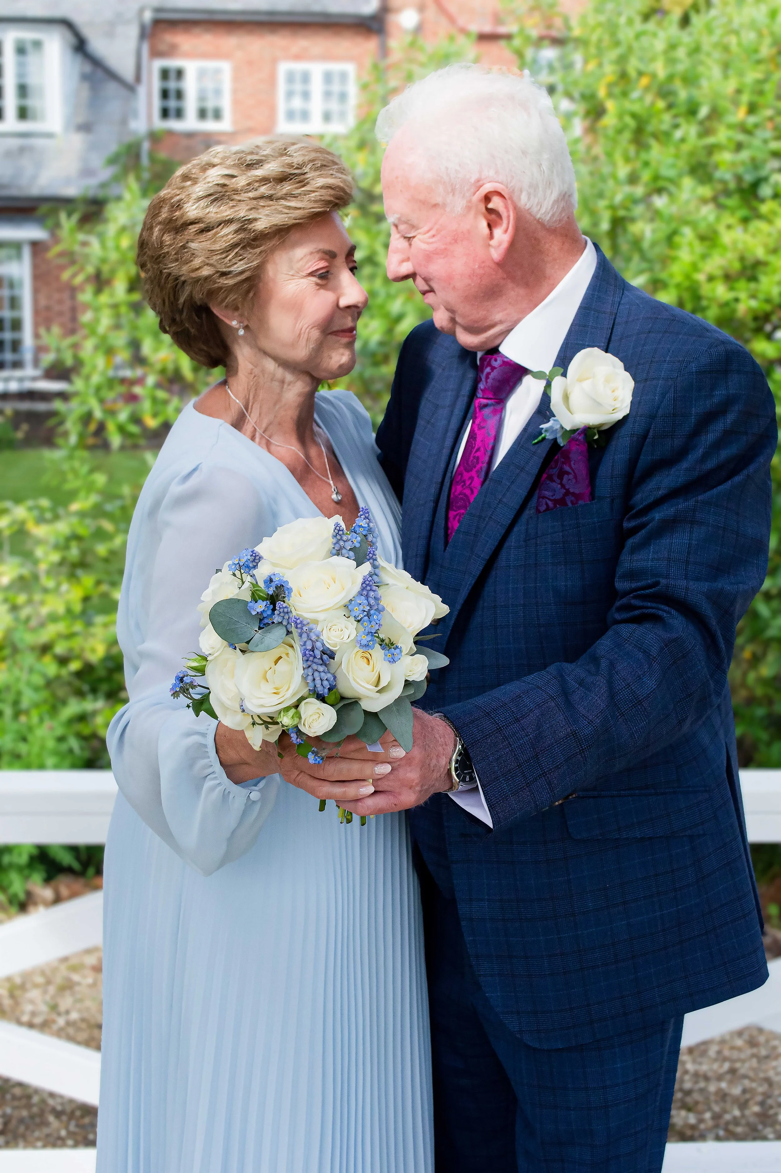 An elderly man and woman embrace at a wedding, the man in a navy suit with a white boutonniere and the woman in light blue holding a bouquet of white roses and blue flowers at The mill house hotel