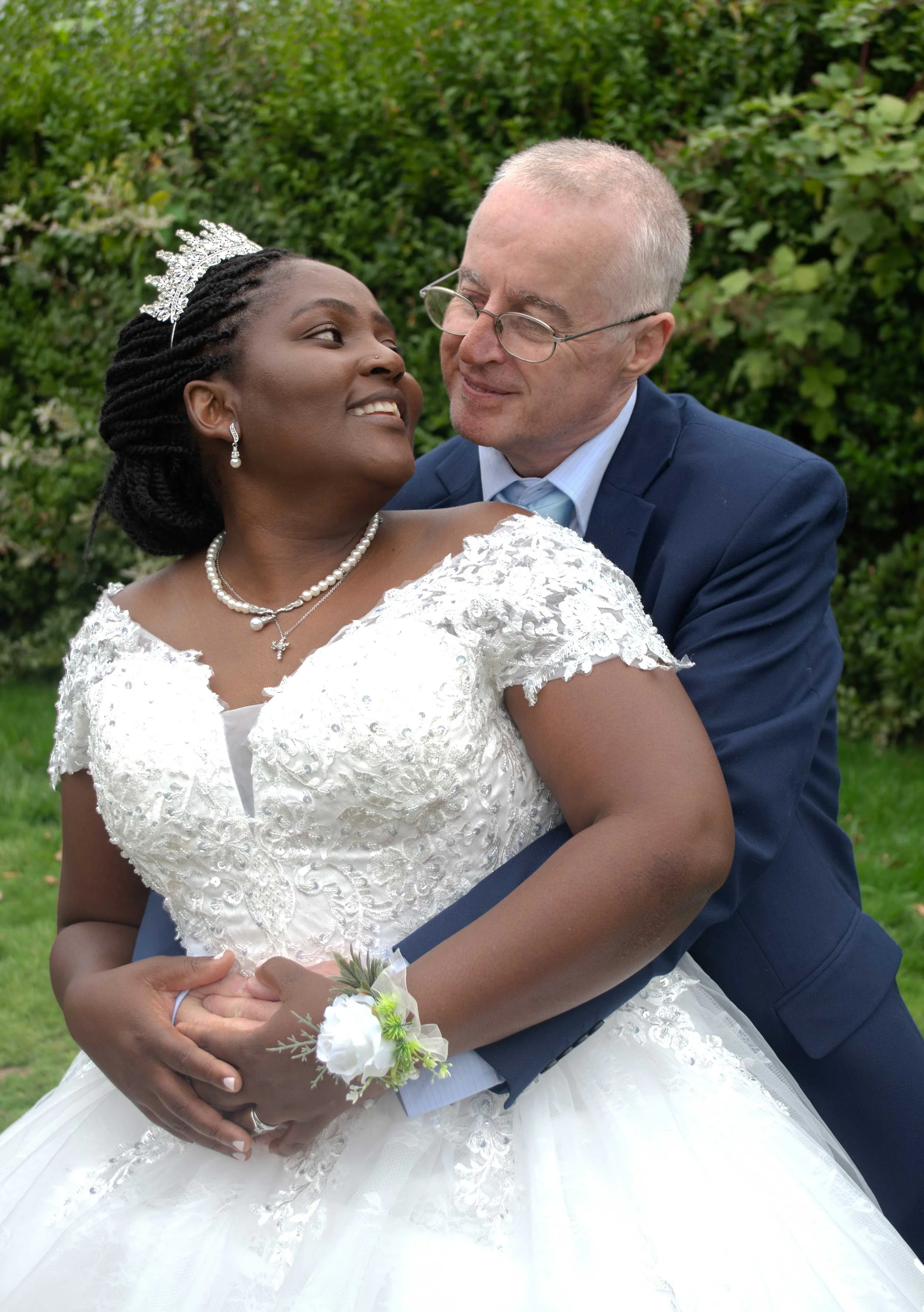A newlywed couple on their wedding day outdoors, with the bride wearing a white lace wedding dress and the groom in a blue suit, sharing a joyful moment at the St Mary Church Camberley