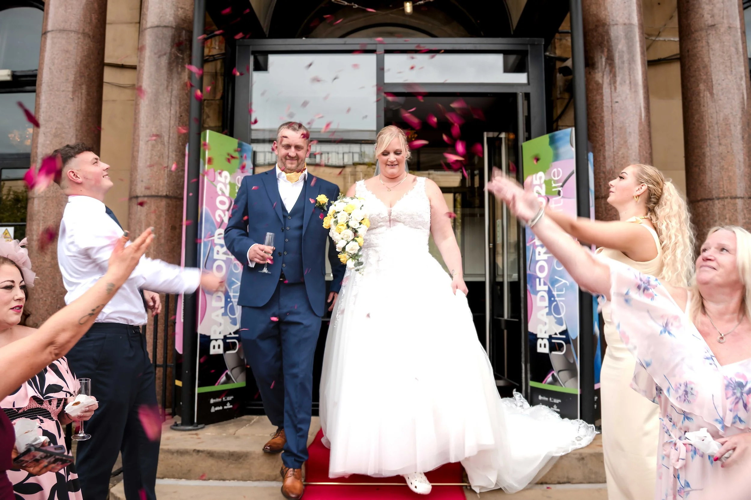 A newlywed couple walks out of a building while friends celebrate throwing flower petals like confetti at Great Victoria Hotel Bradford