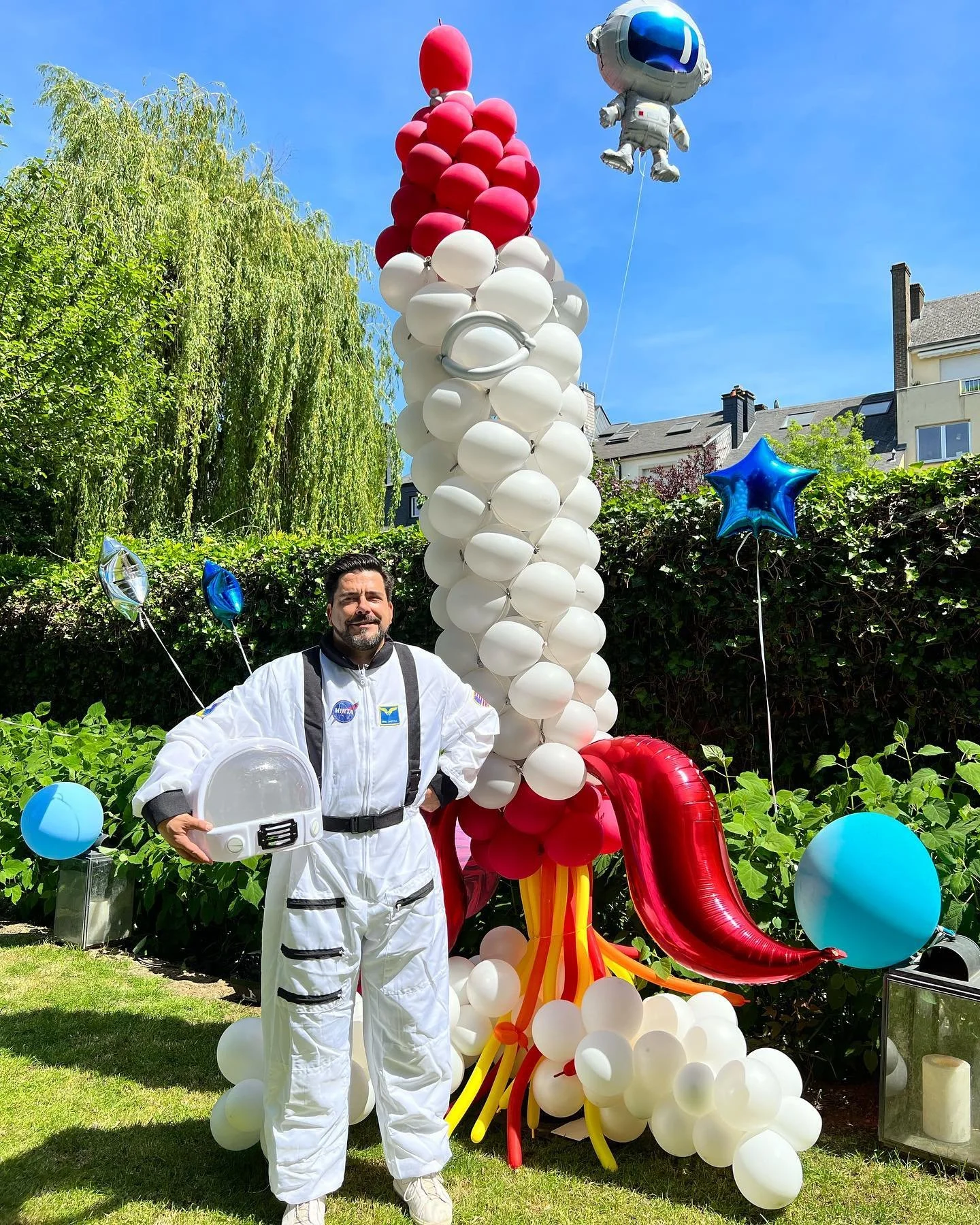 Man dressed in a NASA astronaut suit standing in front of a large balloon display resembling a rocket, with various colorful balloons around, outdoors on a sunny day.