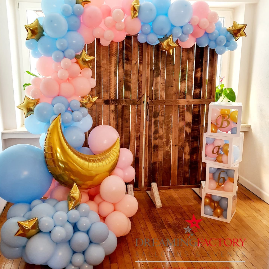 Decorative balloon arch with pink, blue, and gold balloons, including star-shaped balloons and a large gold moon-shaped balloon, set against a wooden backdrop with a white cubical display filled with balloons and gold letters spelling 'Baby' on the side.