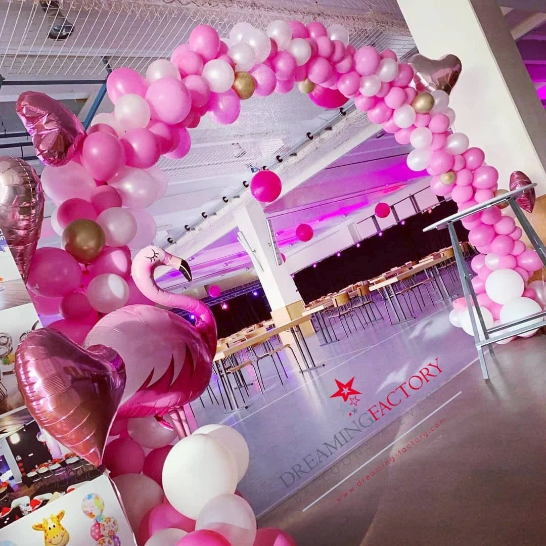 Party decoration with pink and white balloons arranged as an arch, including a large pink flamingo-shaped balloon. The floor has a logo that reads 'DREAMING FACTORY' with a pink star and website link, in an indoor event space with tables and chairs in the background.