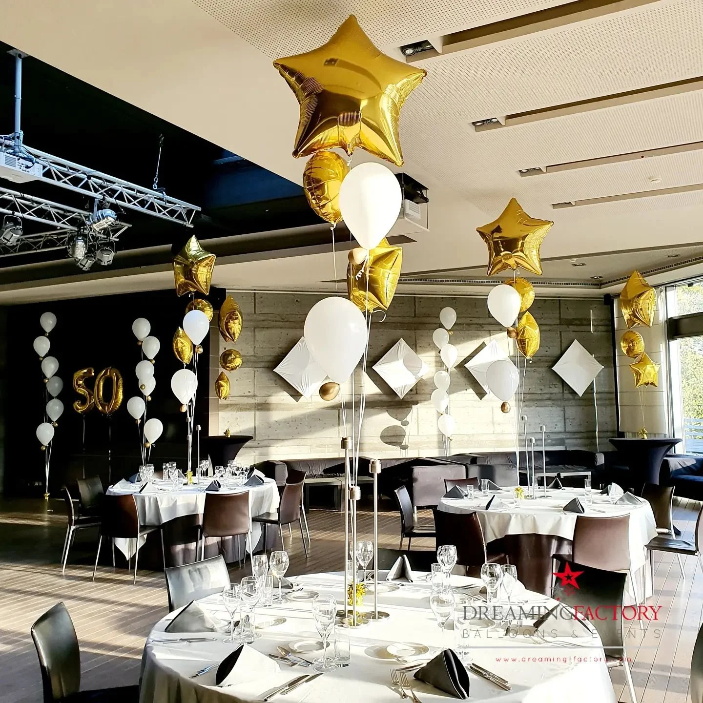 Party hall decorated with white and gold balloons, including star-shaped and round balloons, and gold foil "50" balloons on a black backdrop. The tables are set with white tablecloths, glasses, silverware, and black napkins.