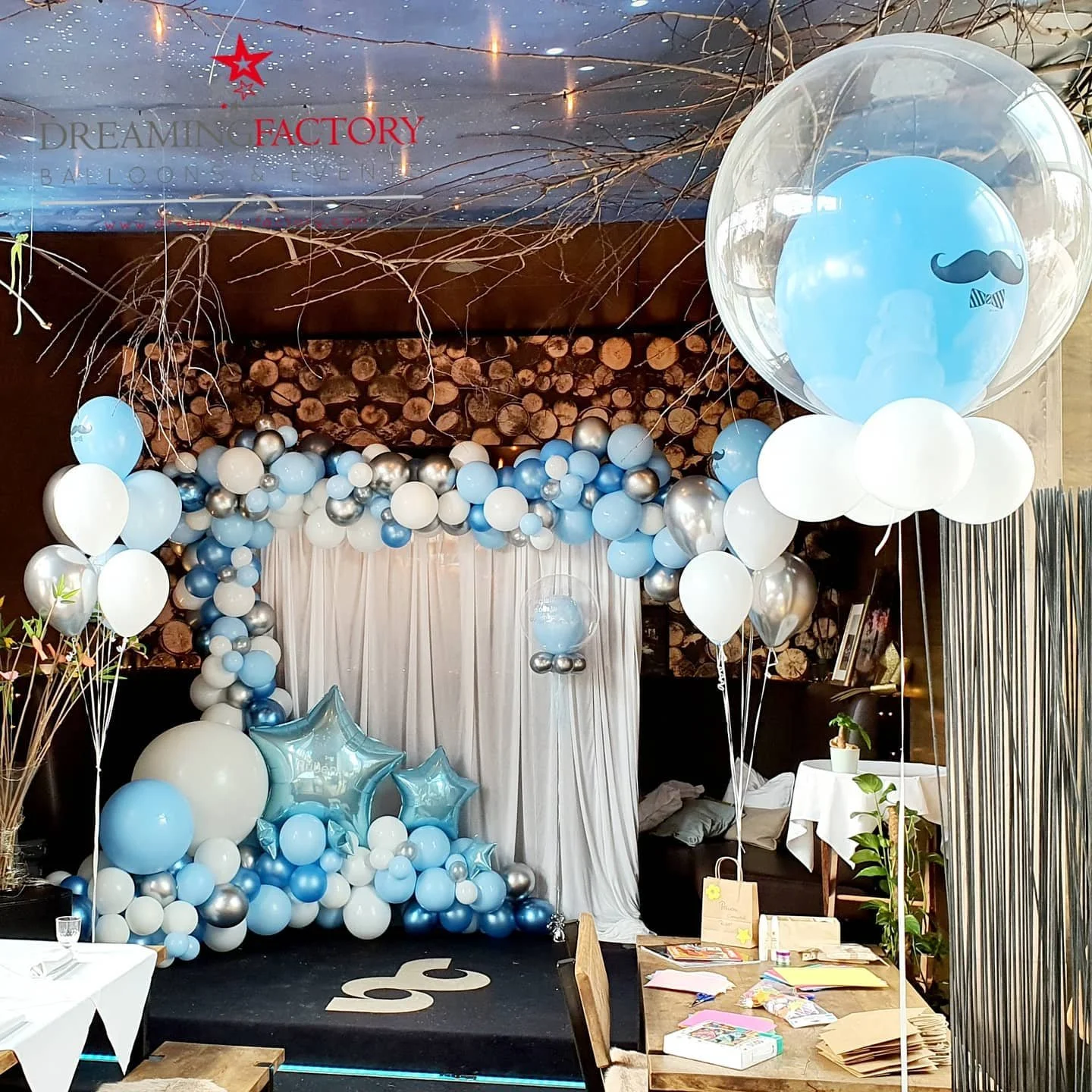Balloon decorations in blue, white, and silver at a party with a balloon arch, star-shaped balloons, and a backdrop. Large helium balloons with mustache and bowtie designs are floating, some with smaller balloons inside.