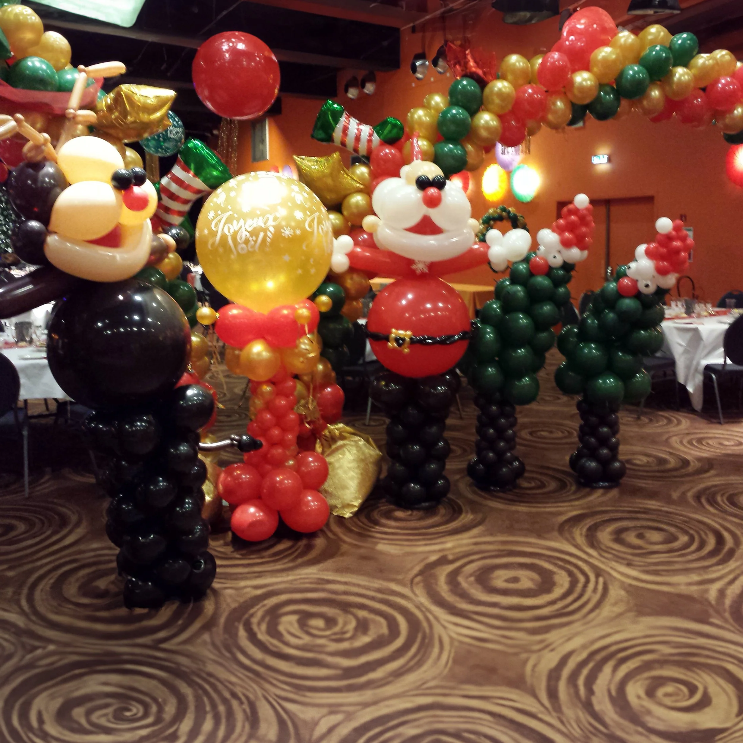 Decorative Christmas balloon sculptures including Santa Claus, Mickey Mouse, and Christmas trees made of balloons, with colorful balloon garlands overhead in a festive party setting.