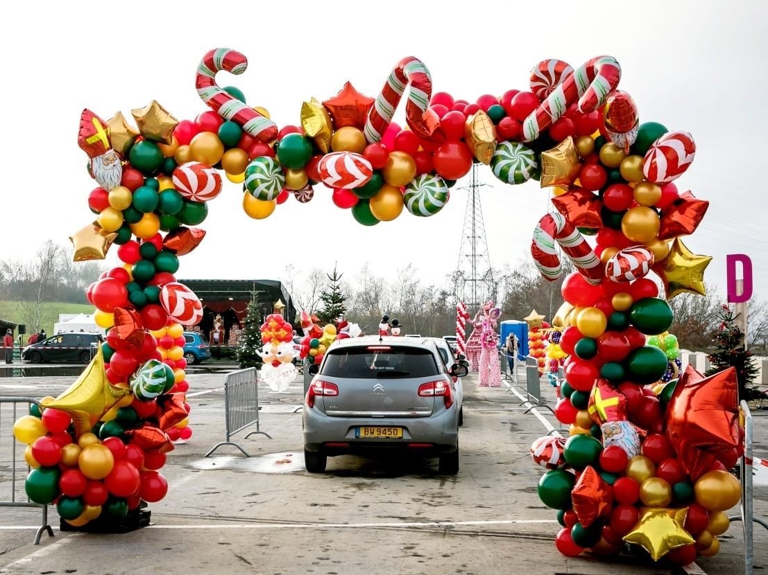 Colorful holiday balloon arch with red, green, gold, and white balloons, decorated with Christmas-themed balloons, at an outdoor event parking lot.