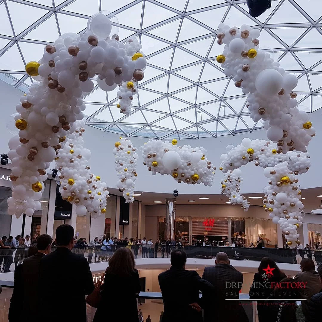 Large white balloon display with gold and copper-colored balloons inside in a shopping mall ceiling area, with people seated and standing below.