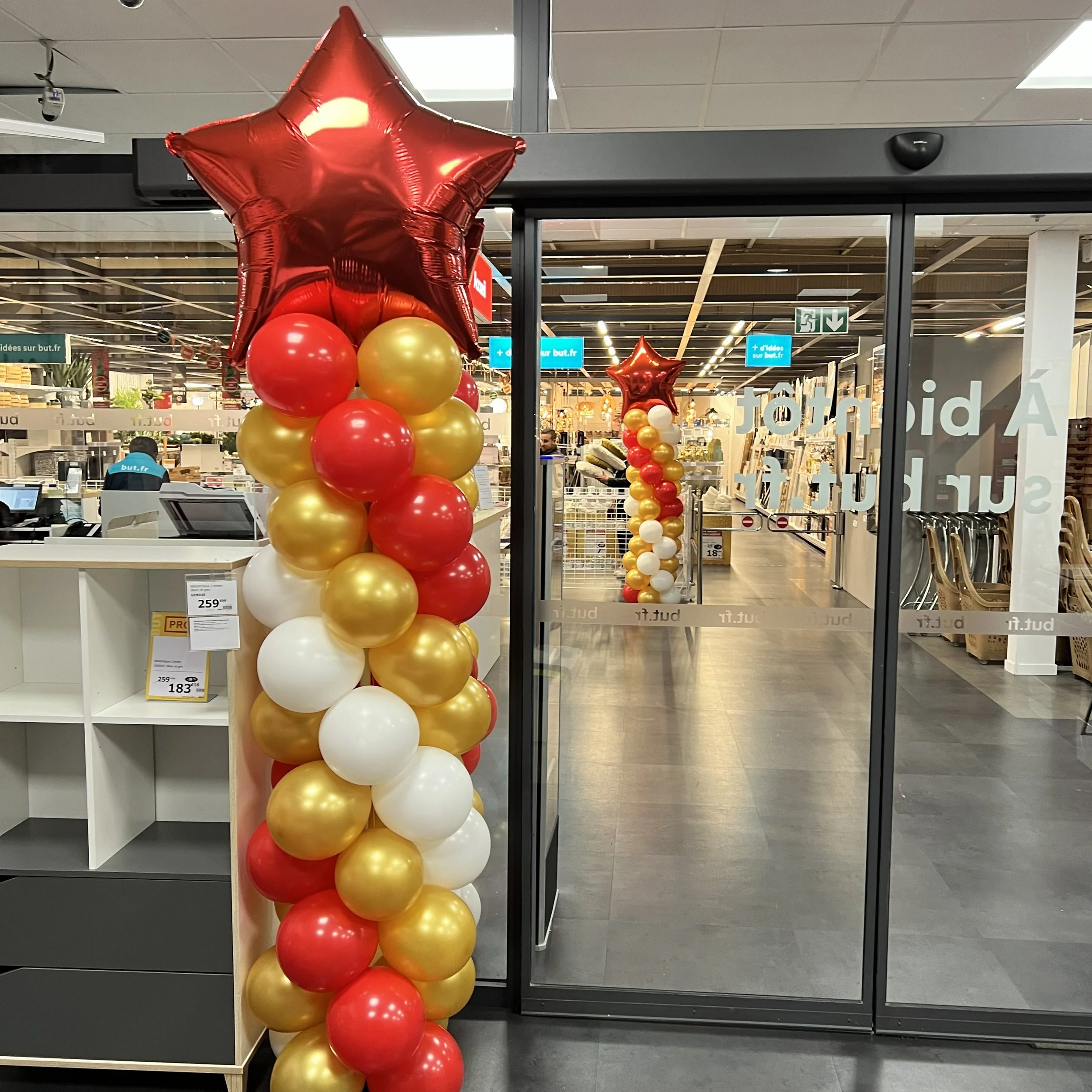 Colorful balloon columns made of pink, green, and blue balloons decorate the entrance of a shopping mall's sidewalk, with parked cars and pedestrians visible.