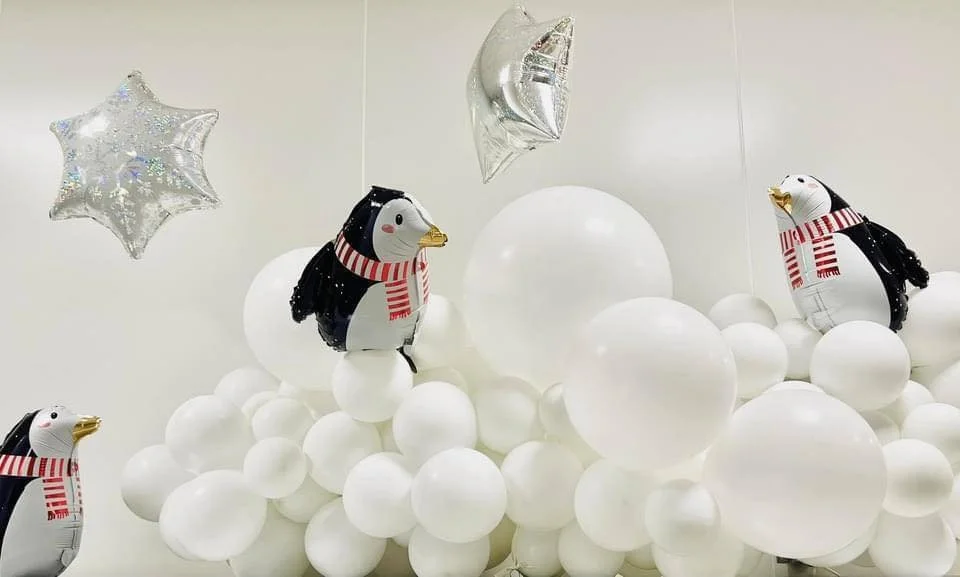 Decorative display with white balloons, penguin-shaped balloons wearing scarves, and silver star-shaped balloons, set up in a festive arrangement.