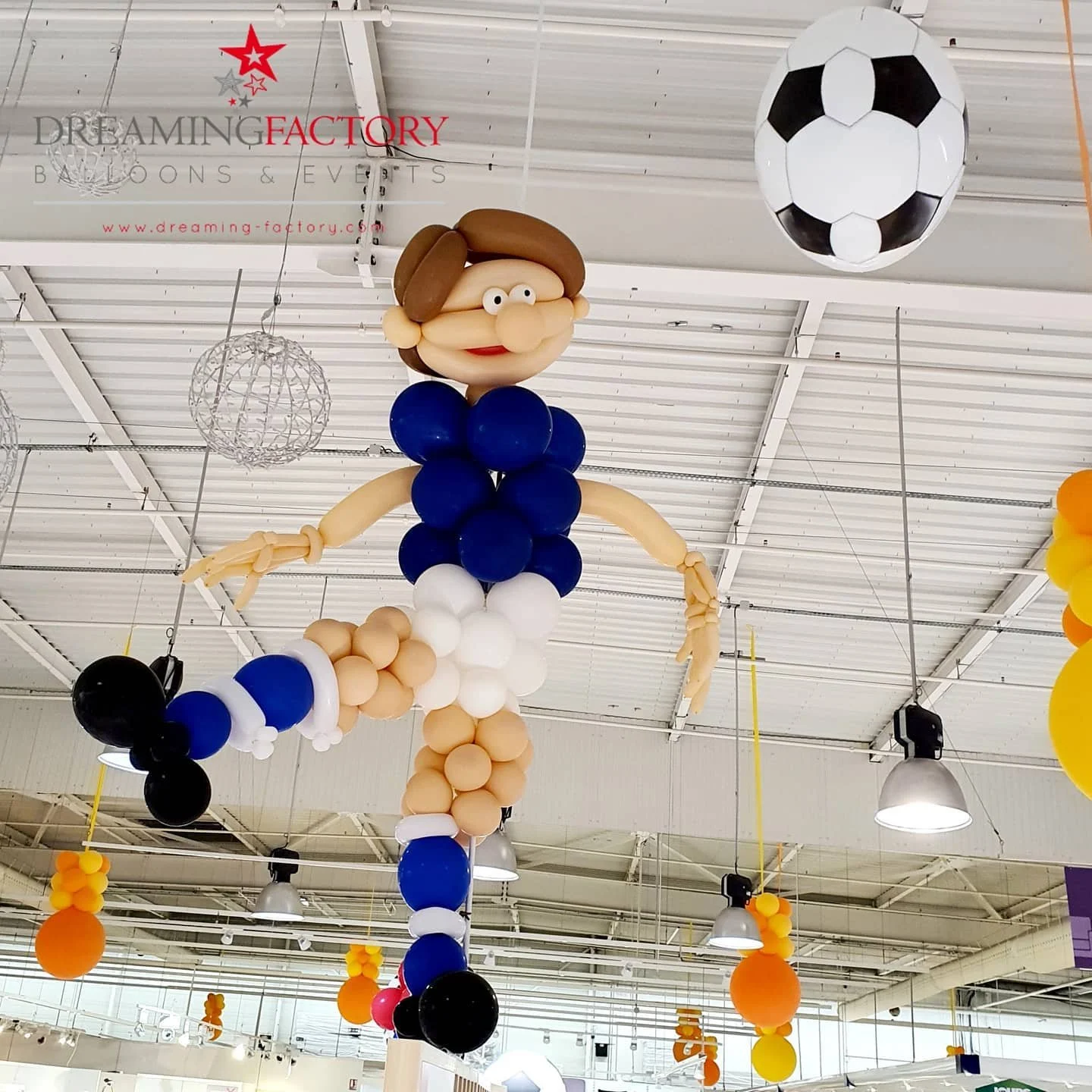 Balloon sculpture of a woman in a blue and white dress with a large head, brown hair, and glasses, hanging from the ceiling, with a soccer ball balloon nearby.