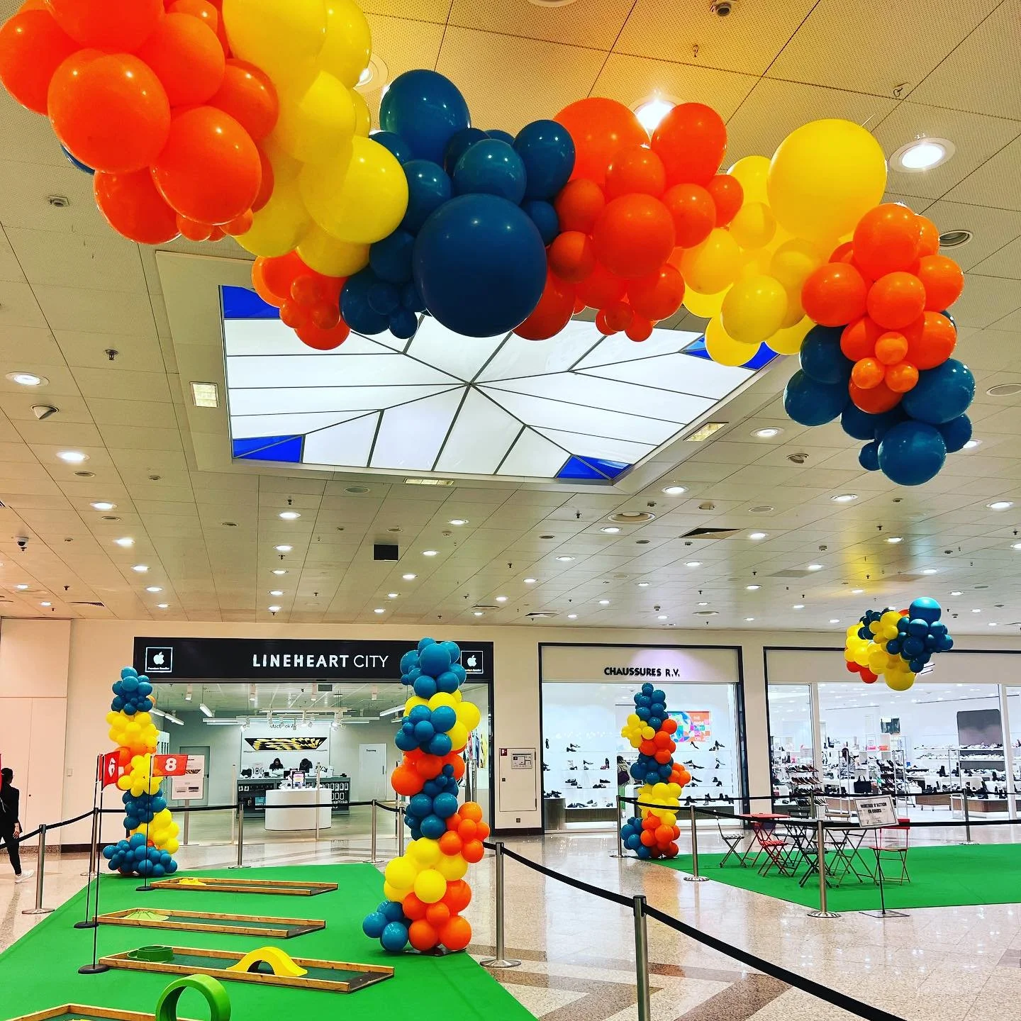 Colorful place decorations with large balloons in blue, yellow, orange, and red, arranged in clusters and arches, set up in a mall for an event or celebration.