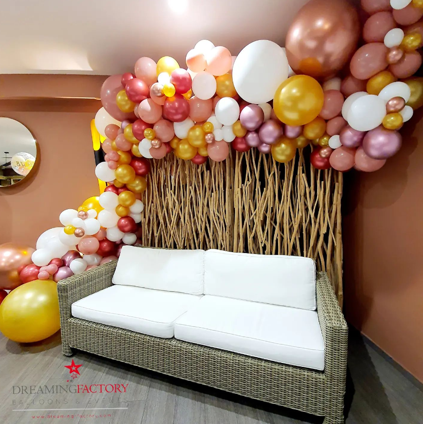 Decorative balloon arrangement with pink, white, gold, and rose gold balloons surrounding a wooden backdrop and a white wicker sofa.