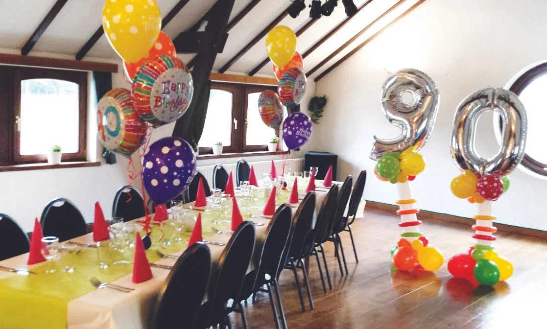 Party decorations with helium balloons, colorful balloons, and silver number balloons displaying 9 and 0 for a birthday celebration in a decorated room with a long table set for a meal.