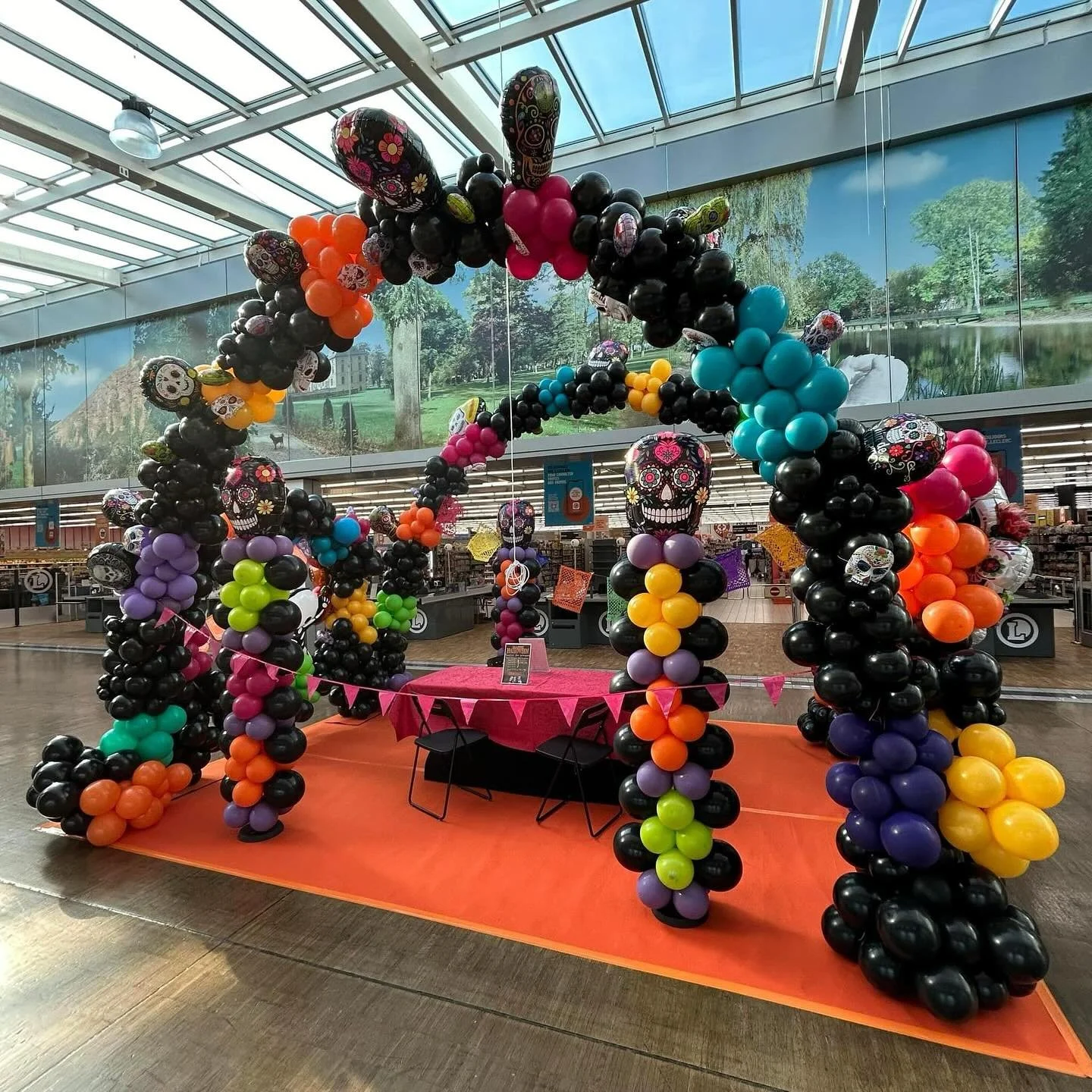Colorful balloon arch and decorations with sugar skull motifs, set up in an indoor space resembling a store or event area, featuring a pink tablecloth and a backdrop of landscape murals.