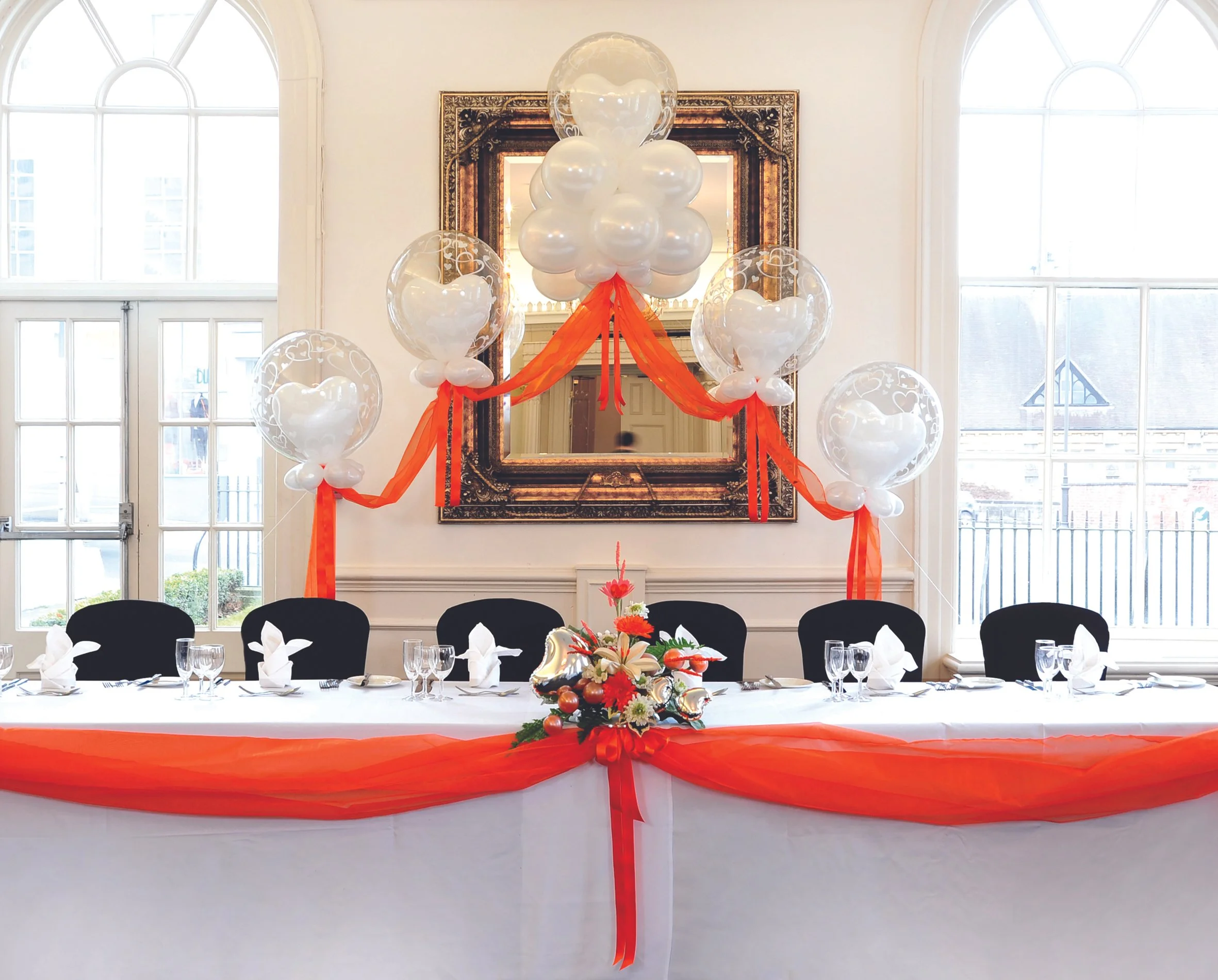 A decorated event table with a white tablecloth, orange sheer fabric, and a floral centerpiece, set for a celebration in a bright room with large windows and a mirror.