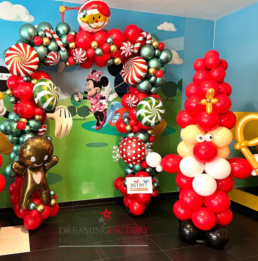 Colorful balloon arch with Christmas theme, featuring a cheerful Santa balloon at the top, surrounded by red, green, gold, and peppermint-patterned balloons. There is a large Rudolph balloon and a Christmas-themed girl balloon, with background decorations of a playful cartoon landscape featuring Minnie Mouse.