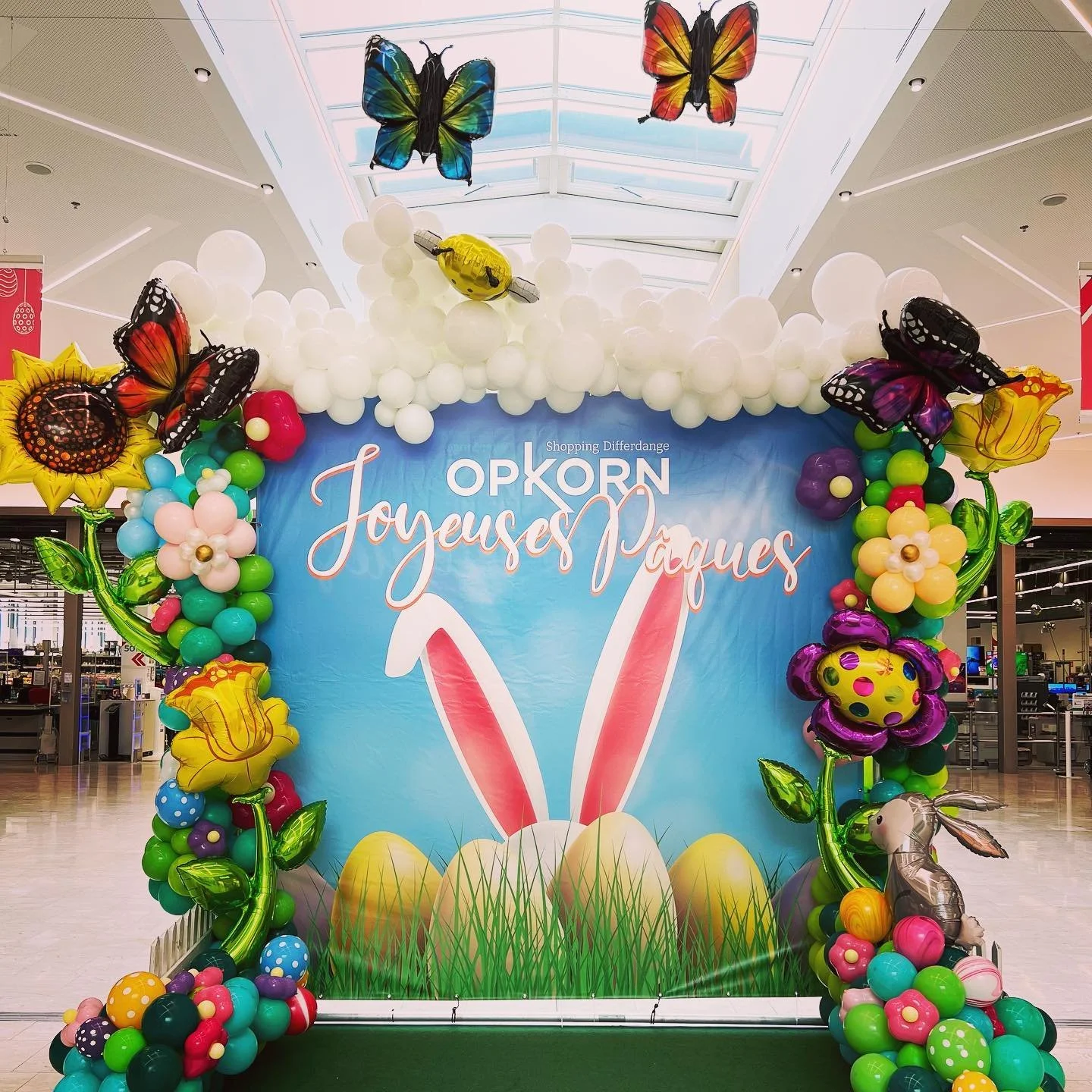 Colorful Easter-themed balloon display with flowers, butterflies, a bunny, and Easter eggs, set in front of a blue backdrop with French Easter greeting text.