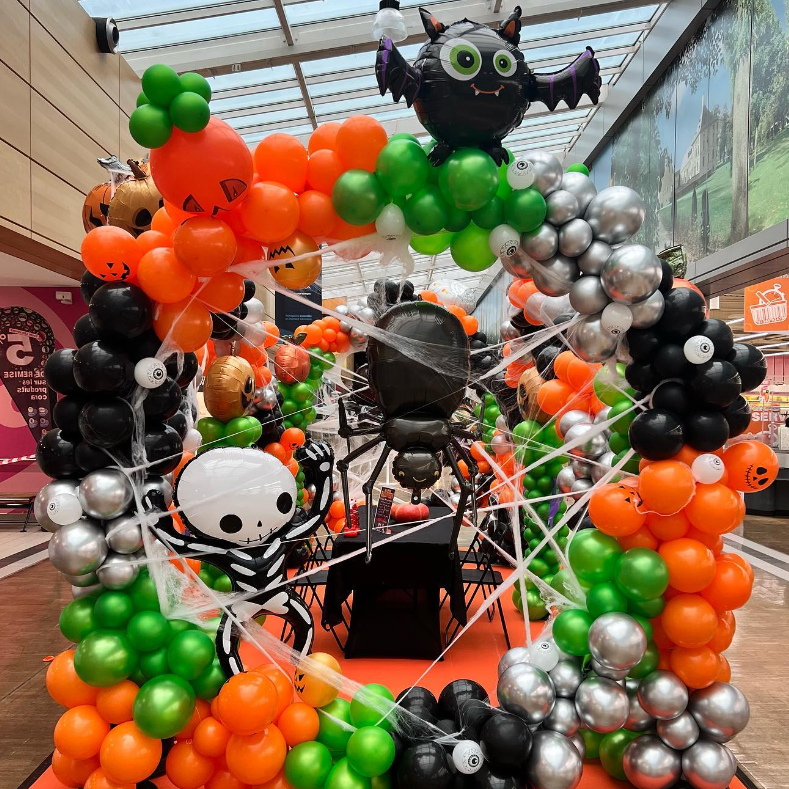 Halloween themed balloon display with orange, black, green, and silver balloons, featuring Halloween characters like a smiling black cat with green eyes and a skull, and spider decorations.