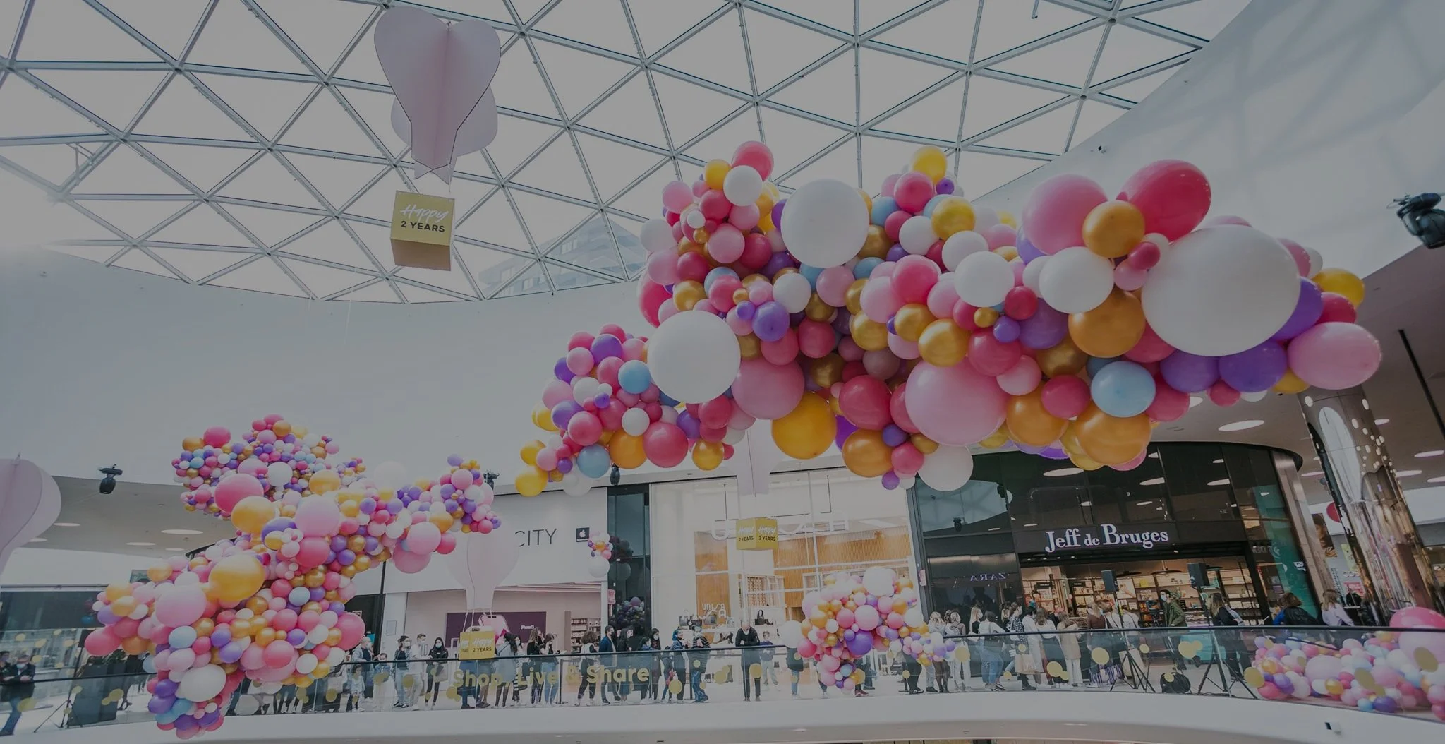 Colorful balloon decorations in a shopping mall with stores and people walking in the background.