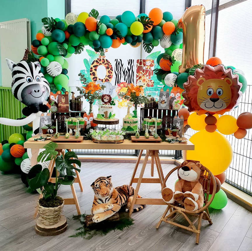 Safari-themed birthday party setup with animal balloons, including a lion, zebra, and tiger, a balloon arch, and themed decorations. A table with treats, flowers, and birthday cakes displays. There is a plush tiger on the floor and a plush lion in a chair, with a large gold number one balloon to the right.