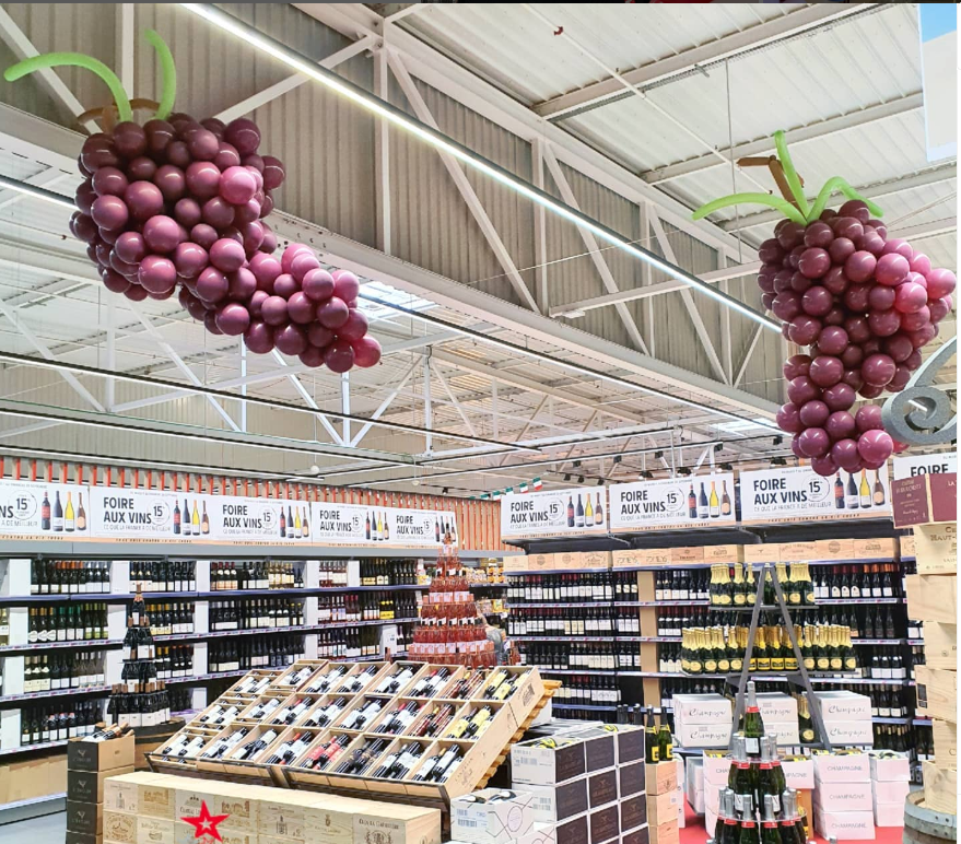 Decorative grape cluster and leaf balloon ceiling decorations inside a store, with wine bottles and wine-related signage on shelves.