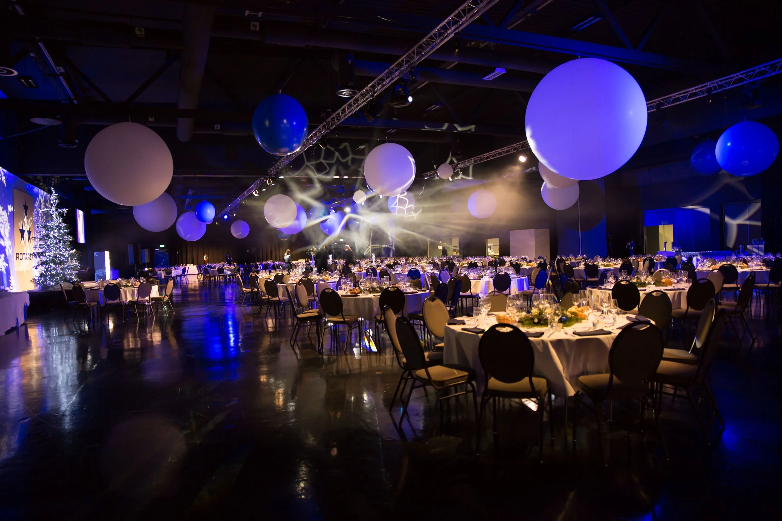 A large banquet hall decorated for a Christmas event, with round tables set with white tablecloths, chairs around each table, a Christmas tree in the corner, large blue and white balloons hanging from the ceiling, and stage lights creating a festive atmosphere.