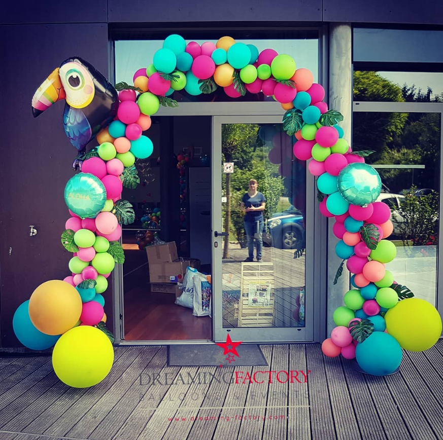 Decorative arch made of colorful balloons and a toucan-shaped balloon at the entrance of a building with glass door and windows, reflecting trees and a person standing outside.