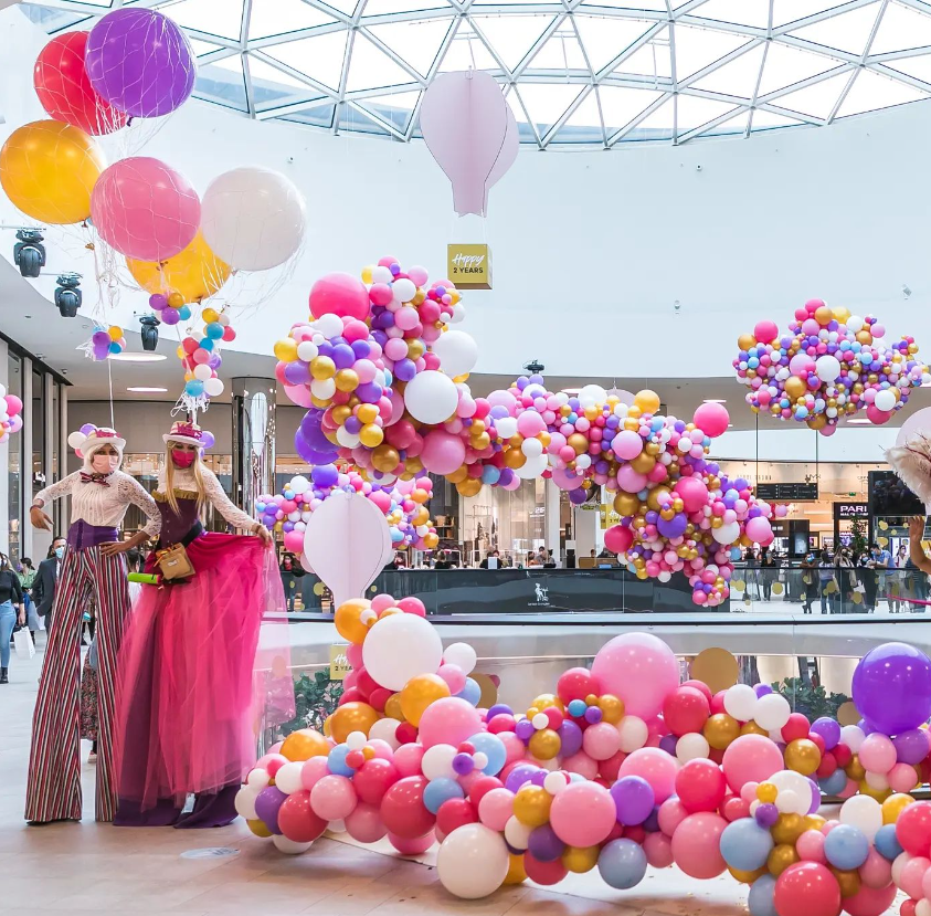 Colorful balloon display inside a shopping mall with mannequins dressed in fashionable clothes and a sign celebrating 2 years.