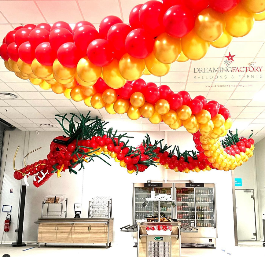 Colorful balloon display featuring red, yellow, and orange balloons arranged in a dragon shape and a winding aerial pattern inside a room with a vending area.