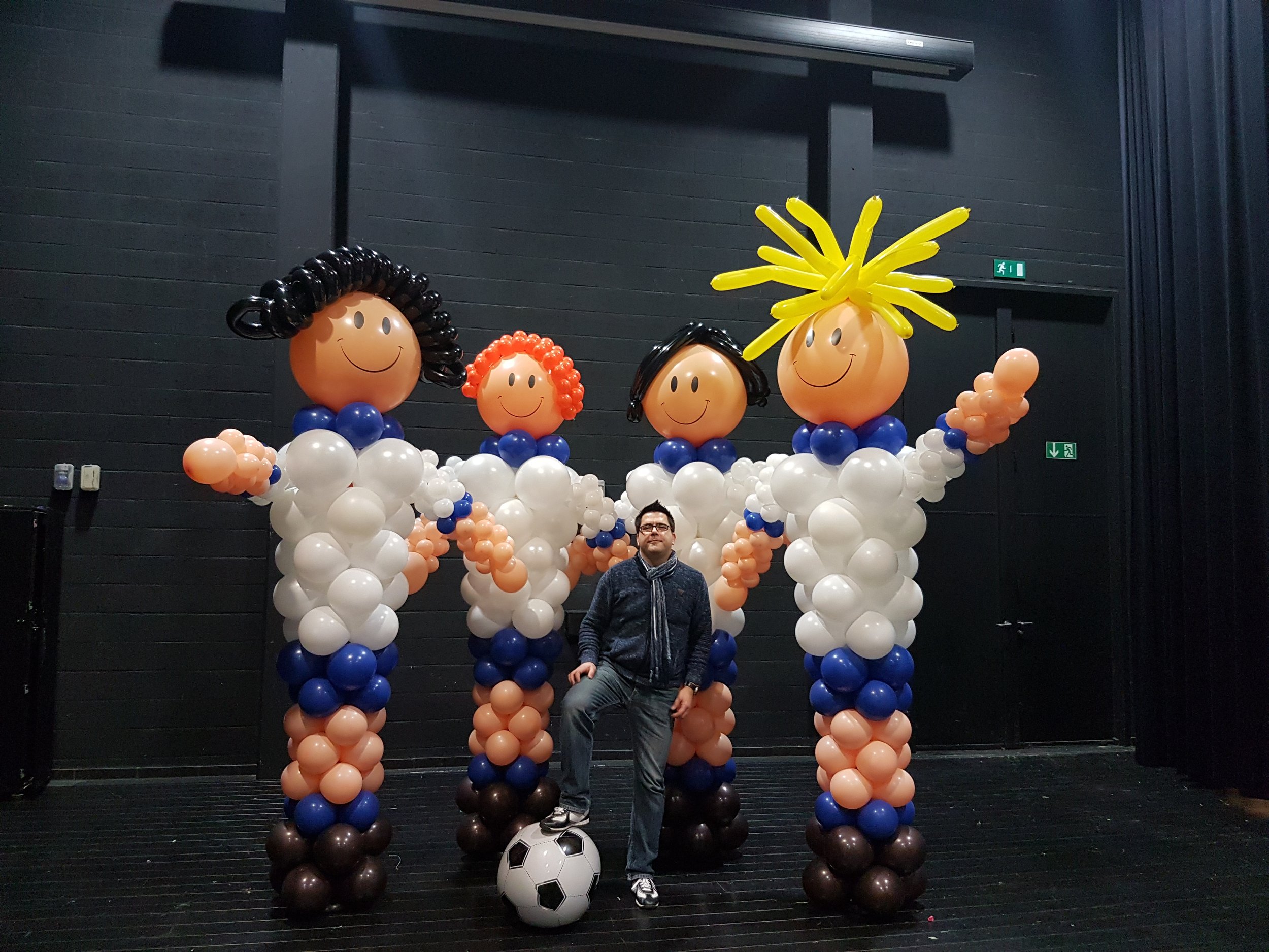 A person standing inside a balloon sculpture of four children with different hairstyles, wearing white shirts with navy blue collars, holding a soccer ball, in an indoor setting with black walls.