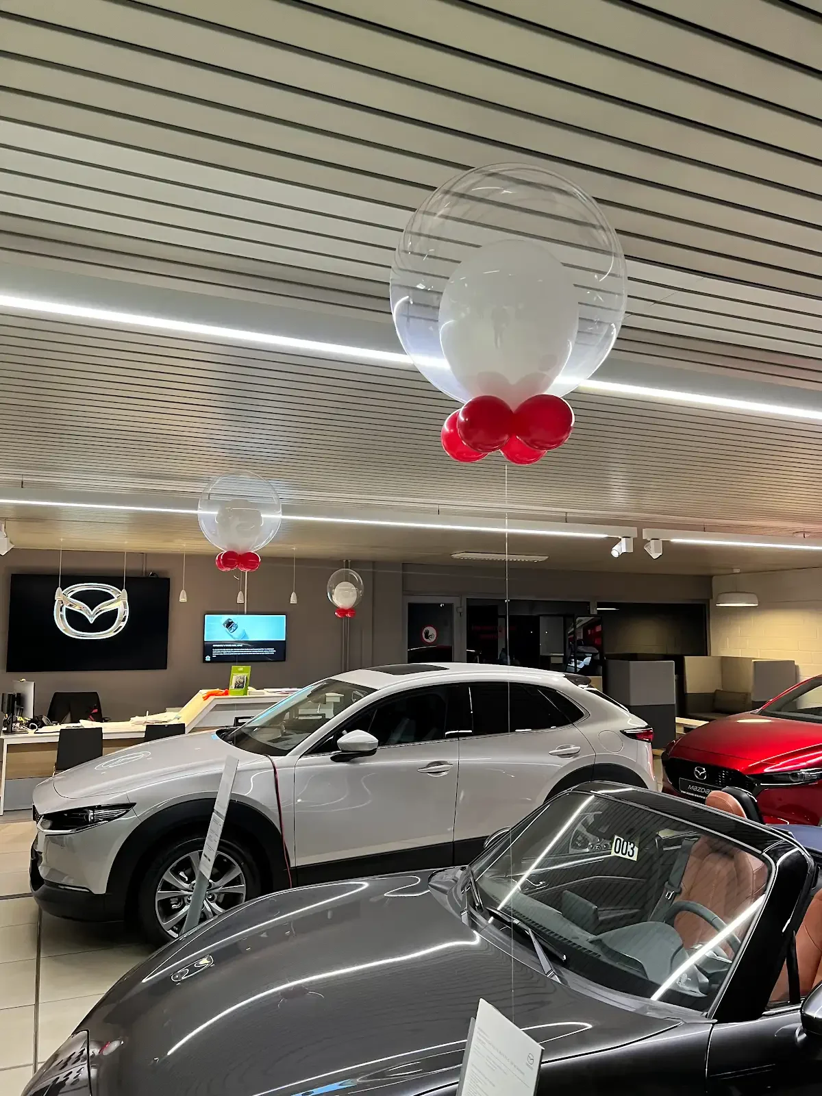 Decorative setup featuring a peacock-style wicker chair surrounded by pastel-colored balloons in pink, purple, and white, with a floral arrangement on a small wooden stand to the left.